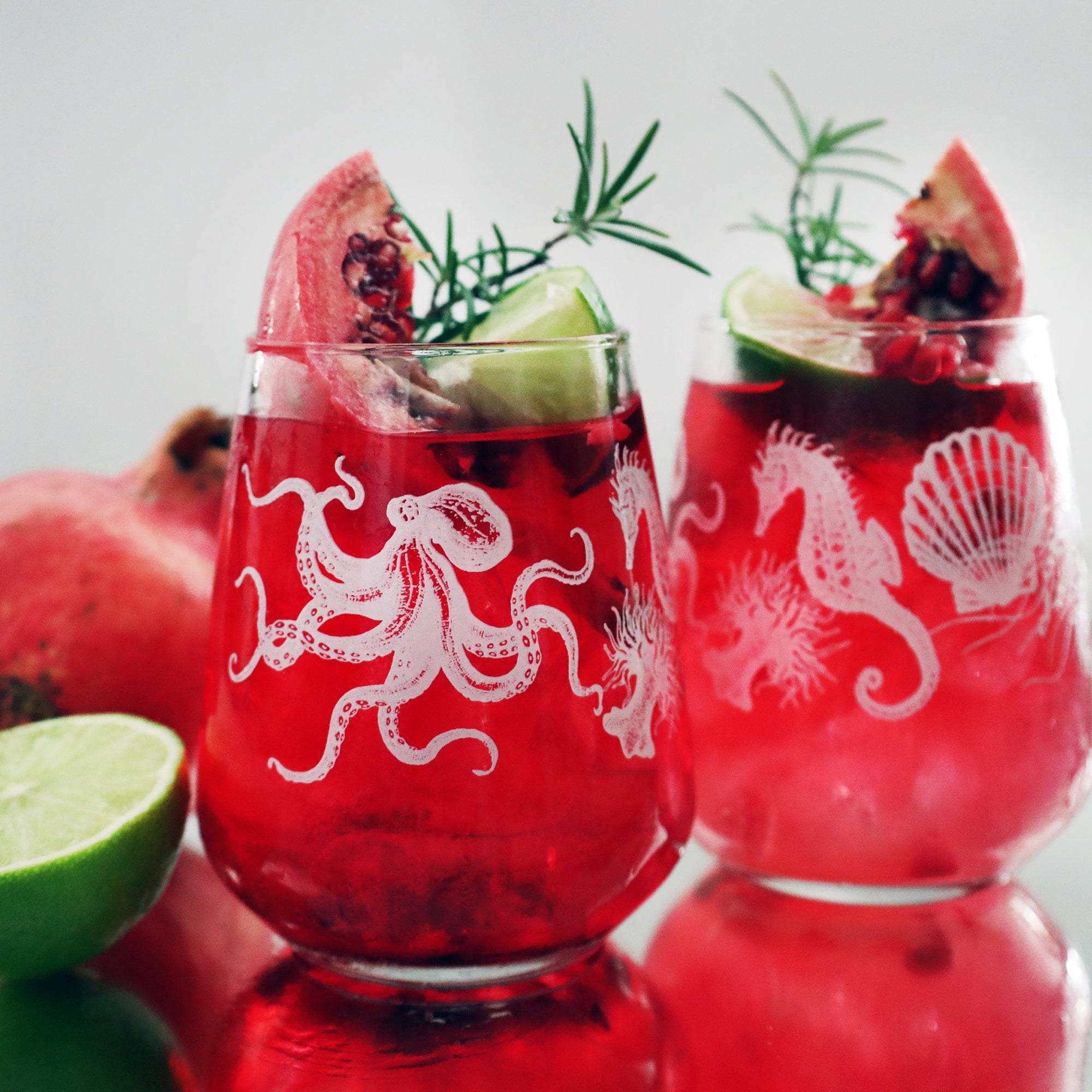 Two glasses filled with a vibrant red drink are decorated with marine-themed designs, featuring an octopus, seahorse, shell, and coral. The drinks are garnished with fresh lime wedges, pomegranate seeds, a sprig of rosemary, and a slice of pomegranate. A few lime and pomegranate fruits are placed in the background, adding to the drink's refreshing and festive presentation. The glasses sit on a reflective surface, giving the image a bright, clean, and elegant appearance.