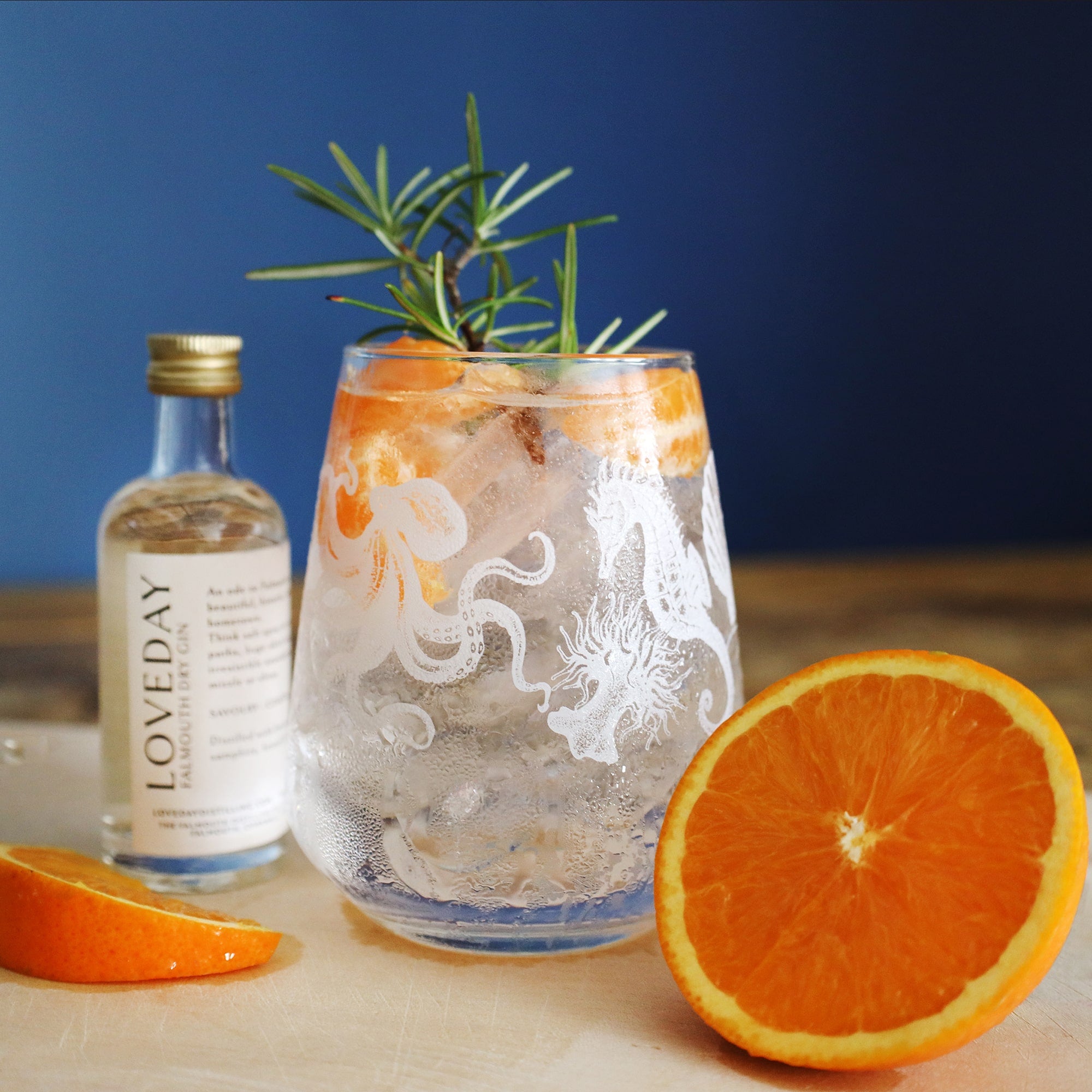 A glass filled with a clear, icy beverage is adorned with detailed marine-themed designs featuring an octopus and seahorse. The drink is garnished with fresh orange slices and a sprig of rosemary, creating a refreshing presentation. Beside the glass, a bottle labeled "Loveday" and a halved orange are displayed on a wooden surface. The deep blue background contrasts with the bright citrus colors, enhancing the drink's vibrant and crisp appeal.