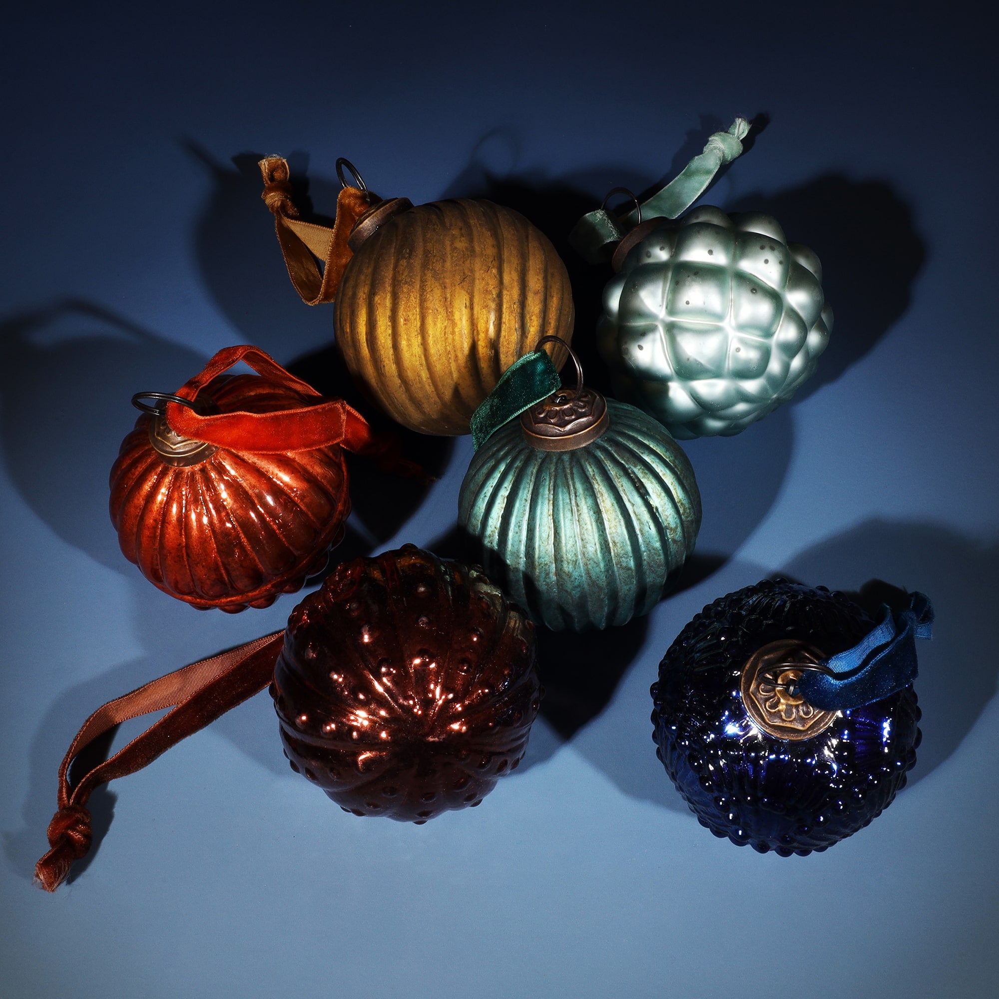 A collection of six vintage-style glass Christmas baubles in various shapes and rich colors, including gold, bronze, teal, blue, and red. Each bauble features a textured surface and is topped with a decorative metal cap, attached to a velvet ribbon for hanging. The arrangement is set against a soft blue background, casting subtle shadows for a festive and elegant look.