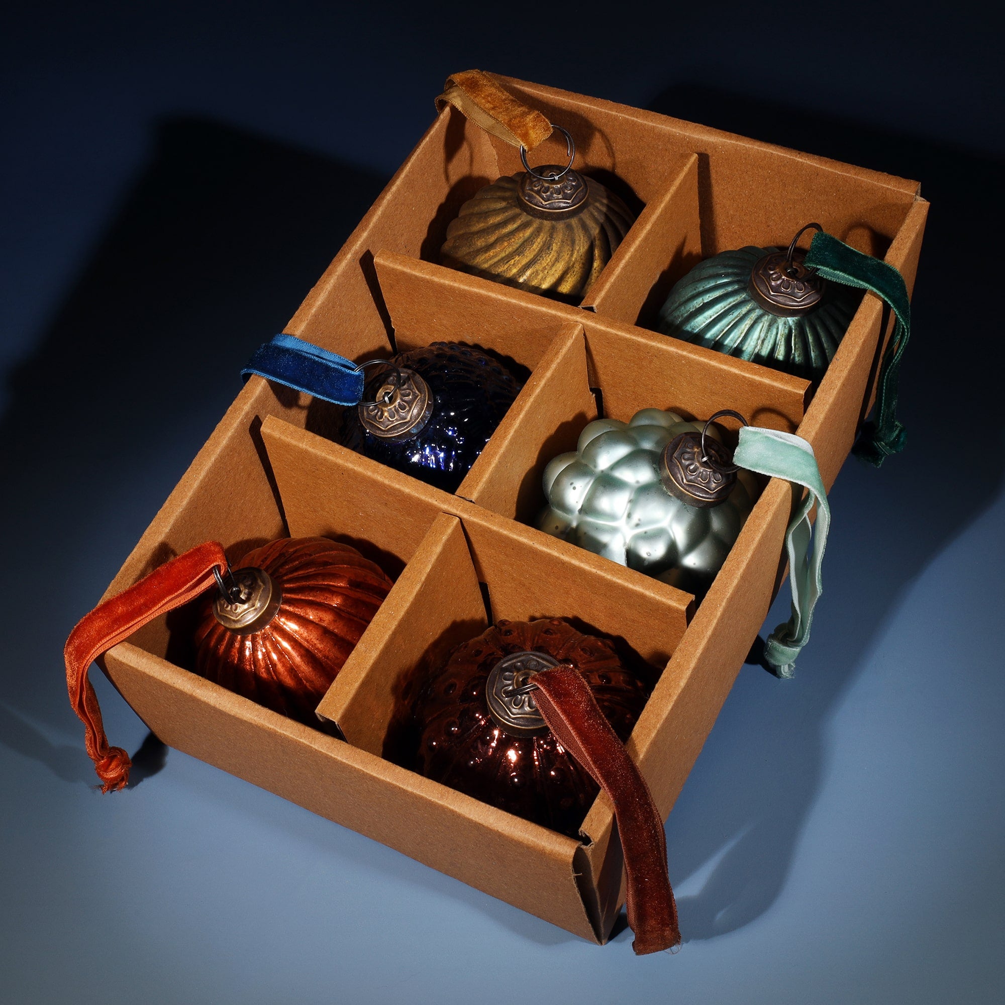 A set of six vintage-style glass Christmas baubles neatly arranged in a brown cardboard box with individual compartments. The baubles come in various shapes and rich colors, including gold, bronze, teal, blue, and red, each with a textured design and a velvet ribbon for hanging. The box provides a protective and organized display, set against a soft blue background.