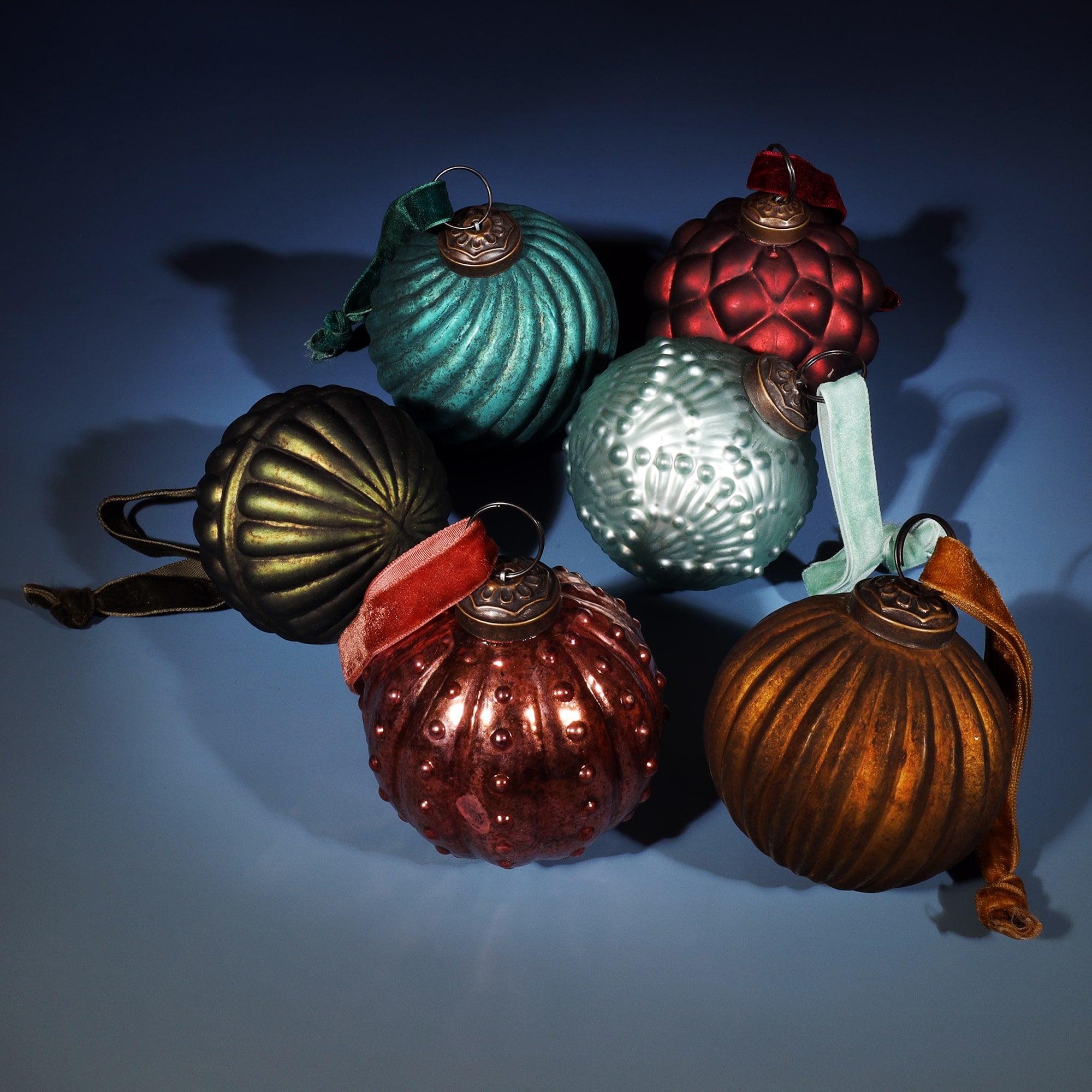 A collection of six vintage-style glass Christmas baubles in various shapes and rich colors, including gold, bronze, teal, red, and light green. Each bauble features a textured design with a decorative metal cap and a velvet ribbon for hanging. The arrangement is set against a soft blue background, casting subtle shadows for a festive and elegant presentation.