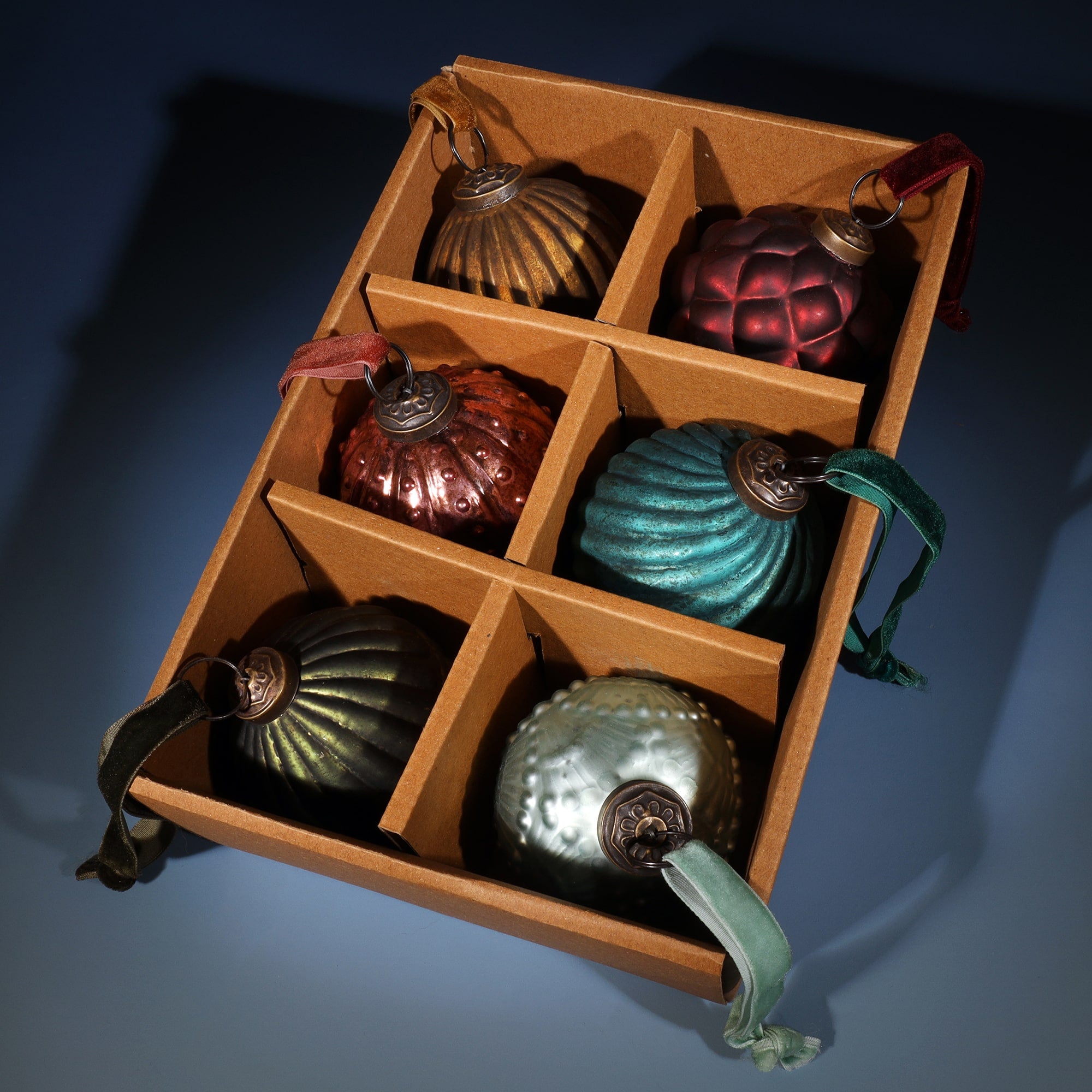 A set of six vintage-style glass Christmas baubles arranged in a brown cardboard box with individual compartments. The baubles feature a variety of shapes and colors, including gold, bronze, teal, and red, with textured designs and velvet ribbons for hanging. The organized presentation is set against a soft blue background, creating a festive and elegant display.