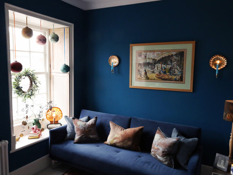 dark teal room scene with christmas baubles hanging in the window and 2 scallop shell brass wall sconces hanging either side of a framed fairground poster, which sits above a navy velvet sofa