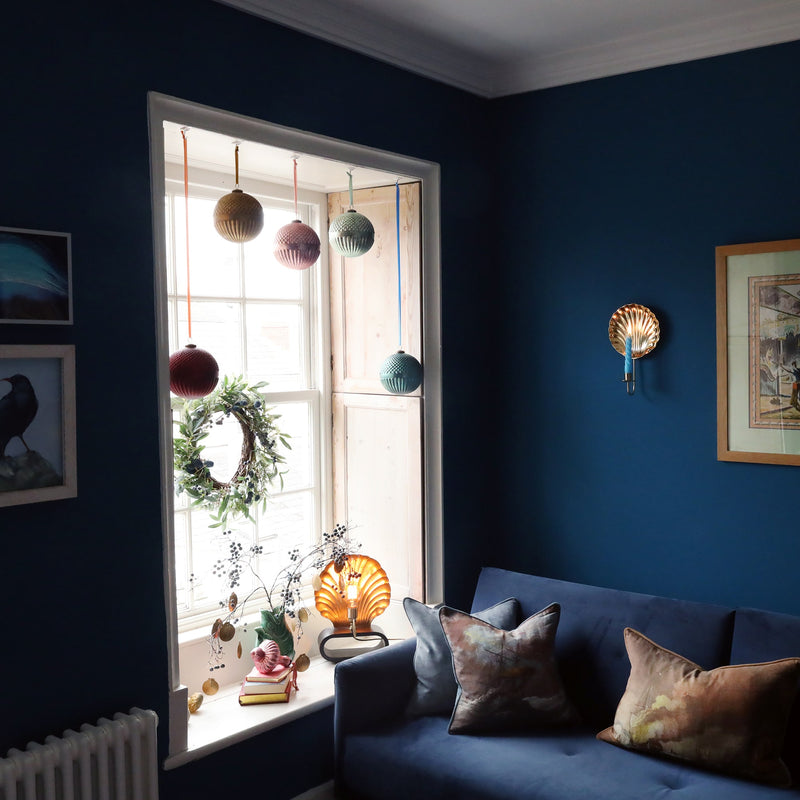 dark teal room scene with christmas baubles hanging in the window and a scallop shell brass wall sconce hanging on the wall above a dark blue velvet sofa 