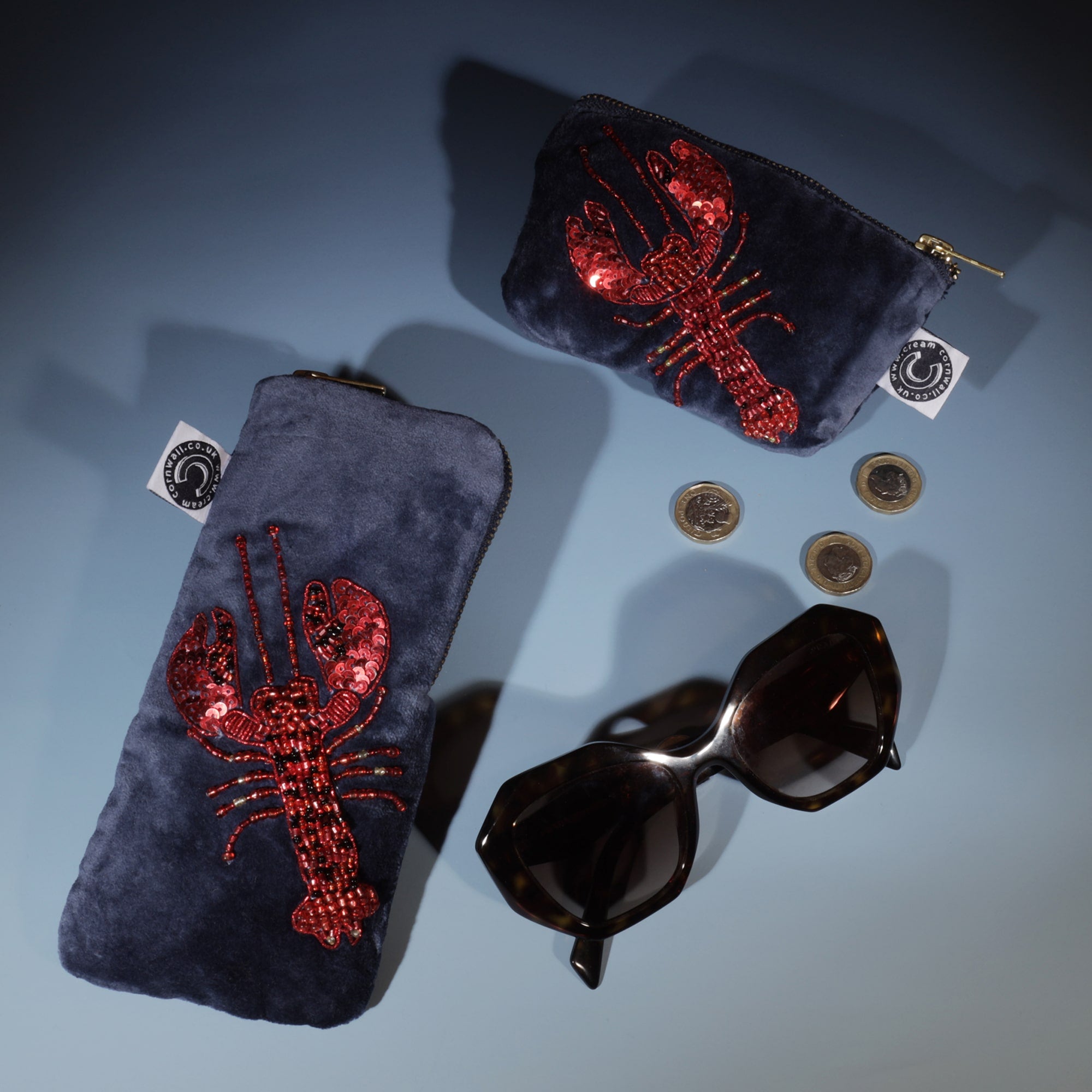 Flat lay of a matching set of velvet accessories featuring red beaded and sequined lobsters on a dark navy background, including a glasses case and coin purse, styled alongside sunglasses and loose coins.