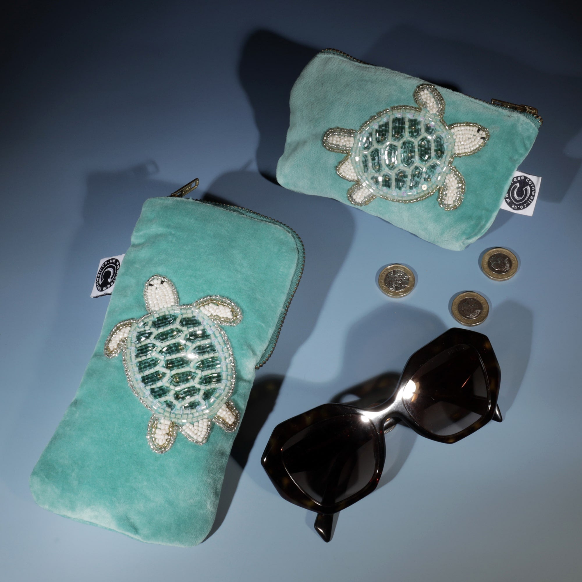 Aqua velvet glasses case and coin purse both with a beaded turtle on them, laid on a graduated blue background with a pair of sunglasses and 3 pound coins.