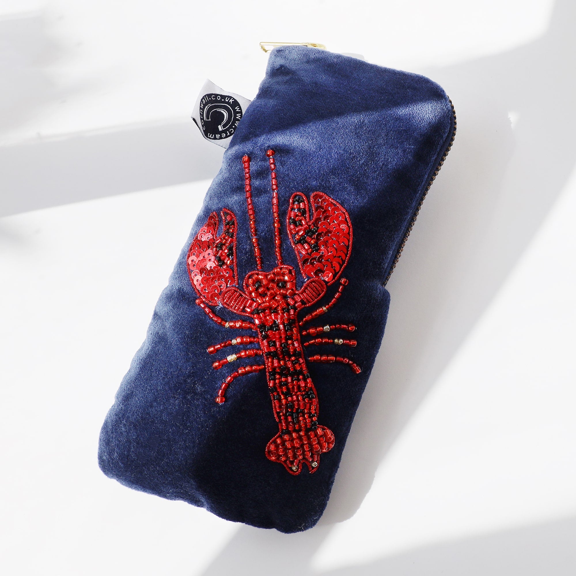 A navy blue velvet glasses case is displayed against a white background with shadows casting soft light. The pouch features a striking design of a red lobster made with sequins and beads, adding texture and sparkle. A small black and white tag is attached to the side, and the pouch is closed with a gold zipper at the top.