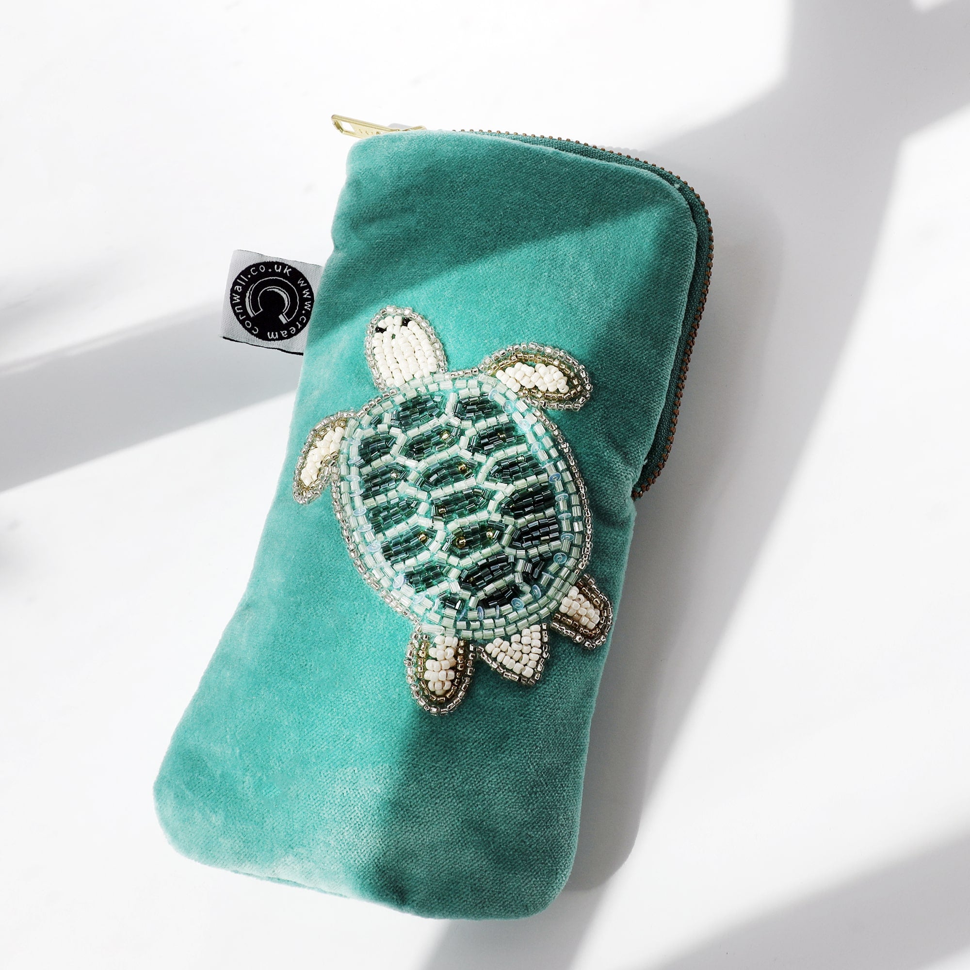 Aqua velvet glasses case with beaded turtle decoration laying on a white background with the sun casting shadows across it.