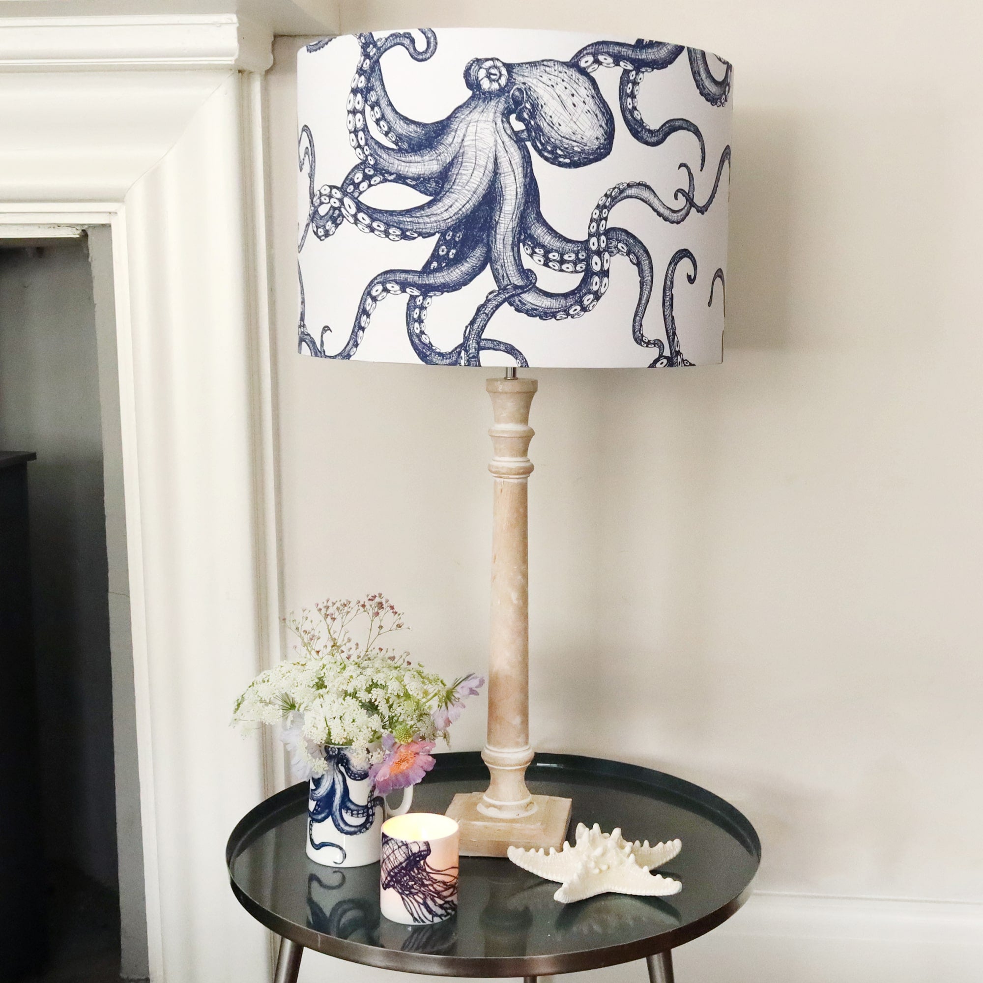 Whitewashed wooden lamp base on side table next to a fireplace with flowers, starfish & flowers and a large 45cm octopus lampshade on top.