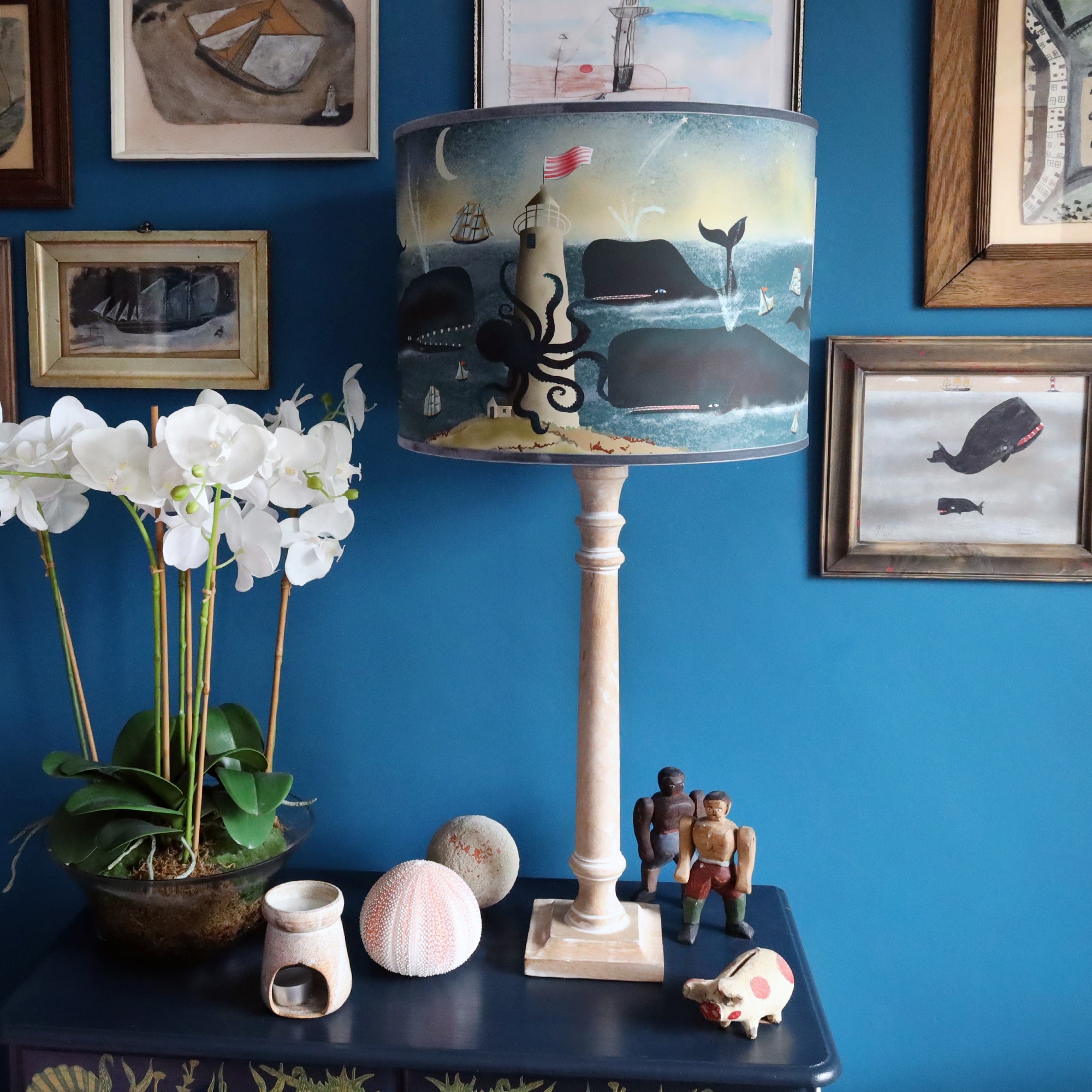 Whitewashed wooden lamp base with whale lampshade, sitting on a set a drawers with naive coastal art and decorative objet around.