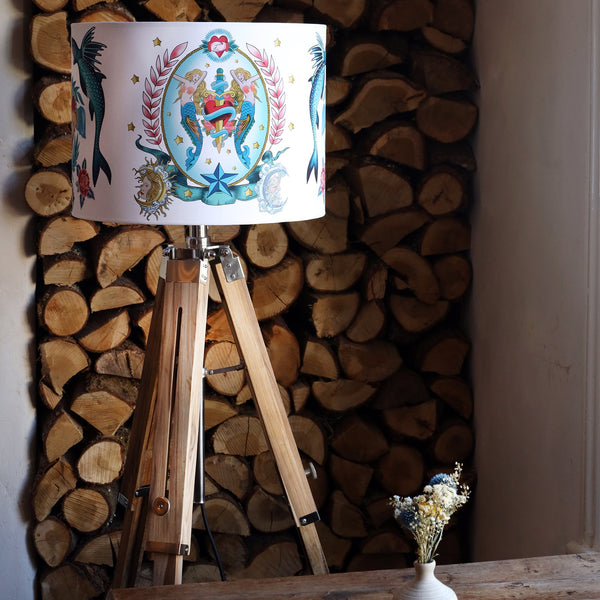 Lampshade on wooden floor tripod with mermaids hearts & daggers design from our Sailor's Story collection. The tripod is standing in front of a stack of cut logs and there is a small wooden table with a vase and dried flowers.