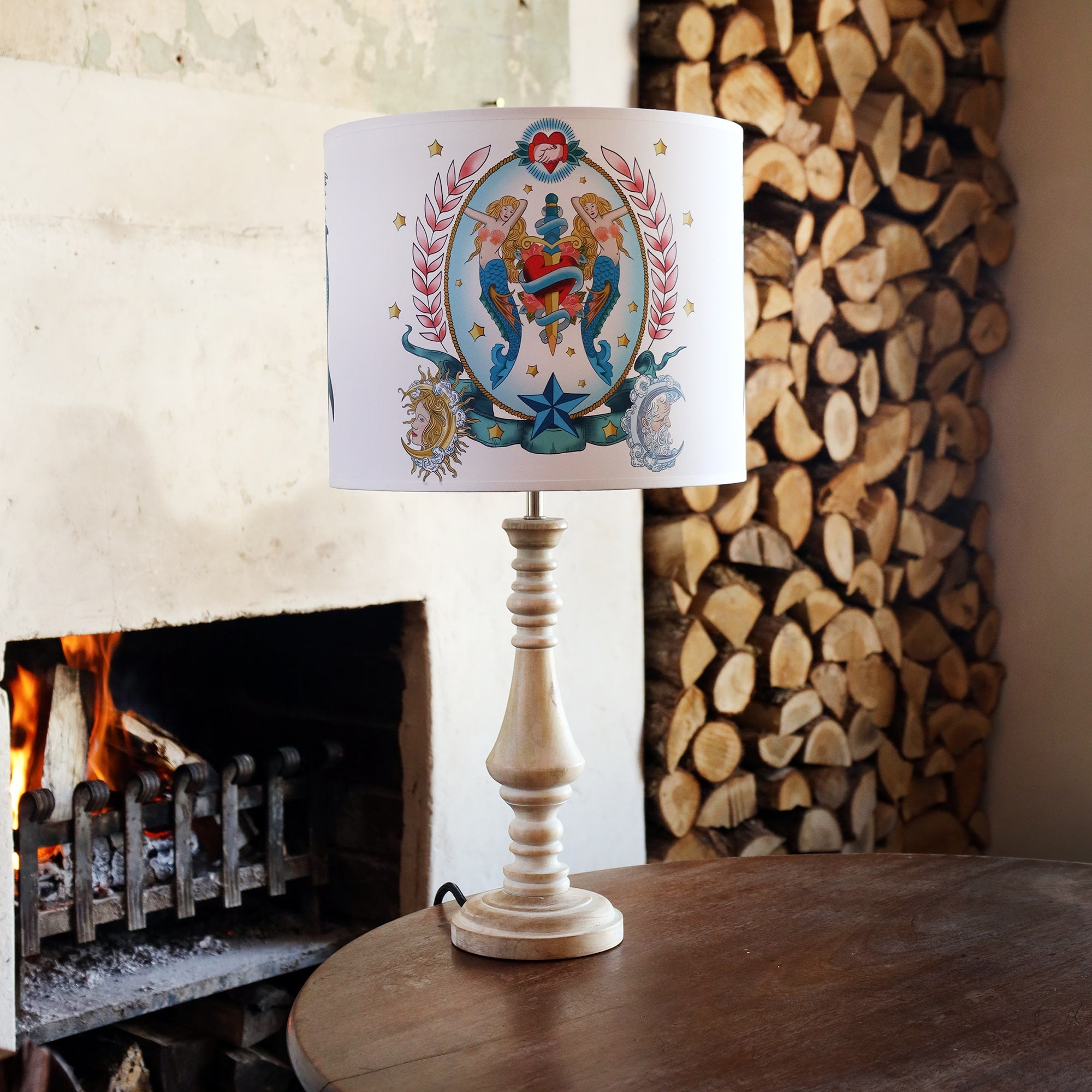 Table lamp on wooden table with a tattoo inspired lampshade with mermaids, heart and dagger design from our Sailor's Story collection. There is a stack of logs in the alcove behind. There is a fire burning in the hearth. 