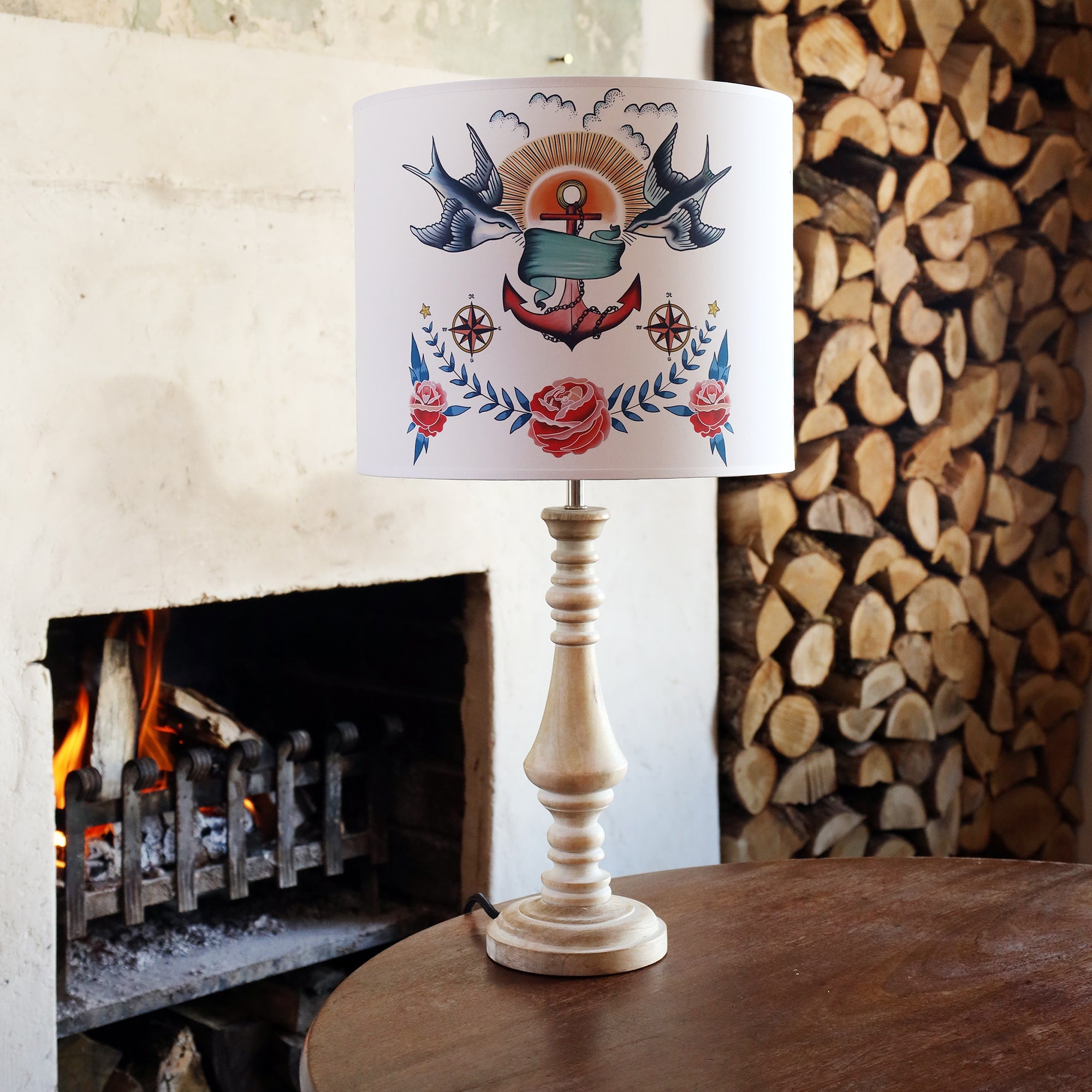 Table lamp on wooden table with a tattoo inspired lampshade with swallows, anchor and roses design from our Sailor's Story collection. There is a stack of logs in the alcove behind. There is a fire burning in the hearth. 