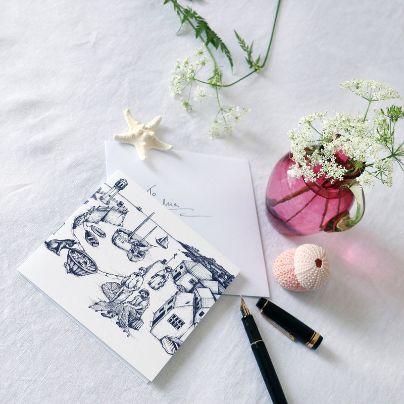greeting card with navy illustration of cornish harbour scene on a white background lying on a white table cloth with a fountain pen, hand written envelope shells and a small cranberry glass jug with wild flowers in 