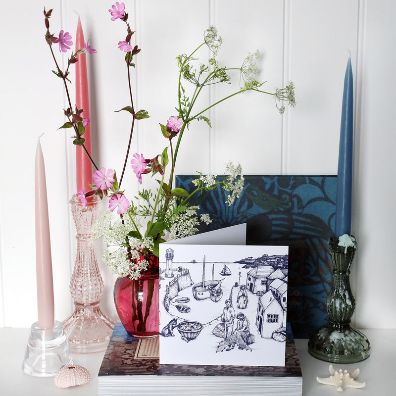 greeting card with navy illustration of cornish harbour scene on a white background on shelf with pink and blue candles in candlesticks and a small cranberry glass jug with wild flowers in
