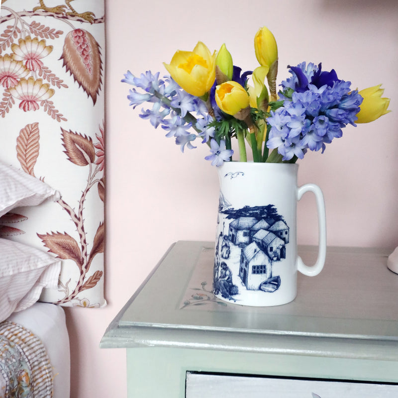 A small white jug decorated with a navy illustration of a harbour scene, filled with yellow daffodils, blue hyacinths and anemones. The jug is on a bedside cabinet with a pink wall behind and you an just see the edge of the headboard and pillows to the left. 