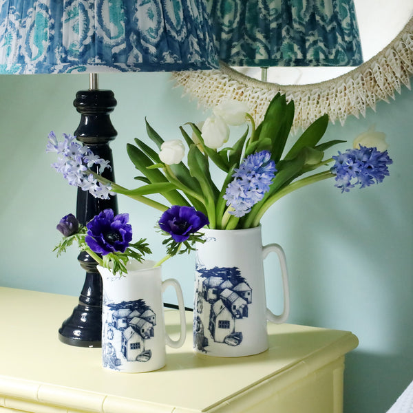 2 white jugs decorated in navy harbour scene illustration filled with blue hyacinths, anemones and white tulips. they are on a yellow set of drawers with a navy lamp base and block printed lampshade. The wall behind is a soft green/blue and you can just see the edge of a mirror decorated in white cut shells. 
