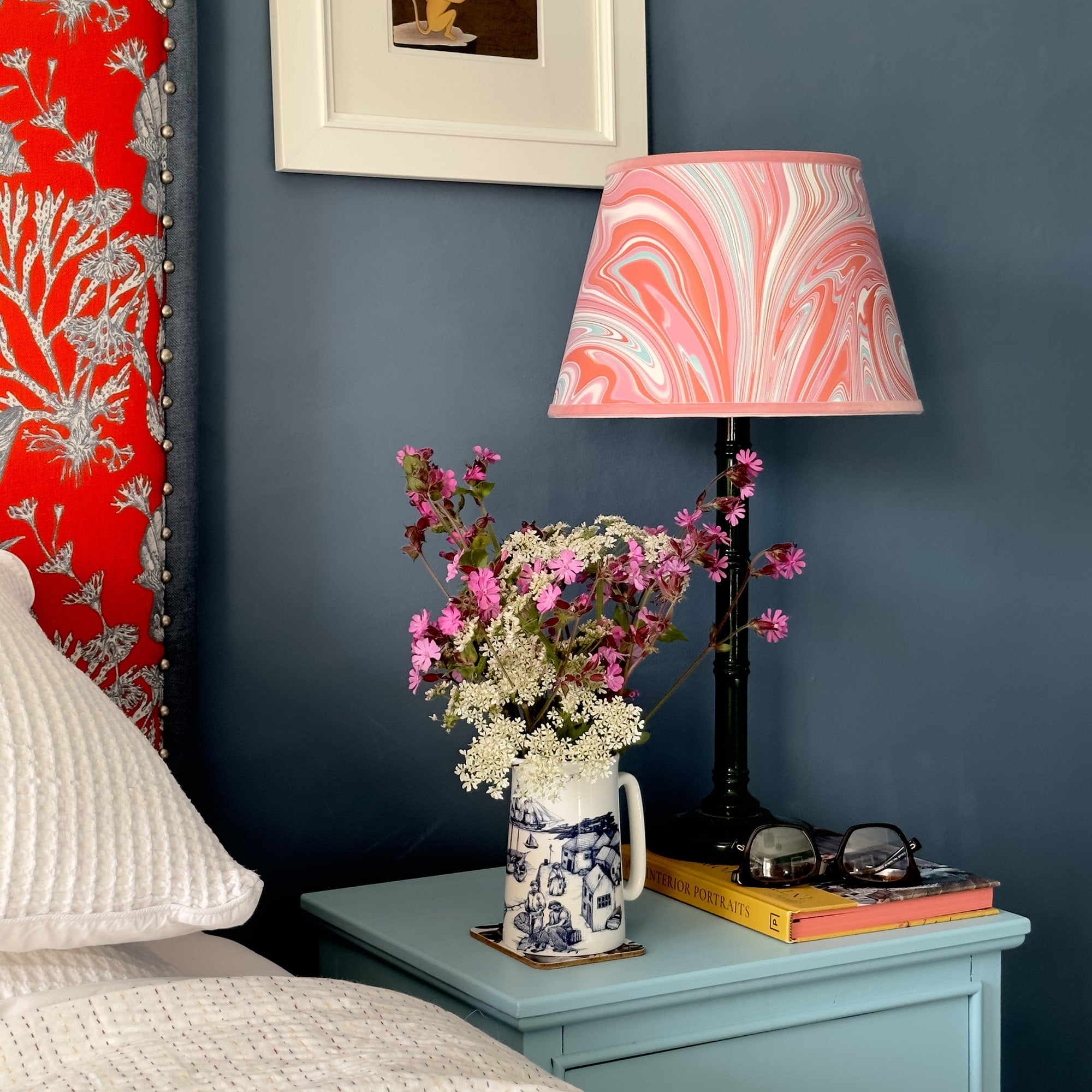 A medium white jug with a navy illustrated harbour scene on it filled with wold flowers in white and pink. This is sitting on pale blue bedside cabinet with a book and reading glasses next to it and a dark green glossy lamp base and an orange marbled effect lampshade. all this is set again a lovely denim blue wall.   