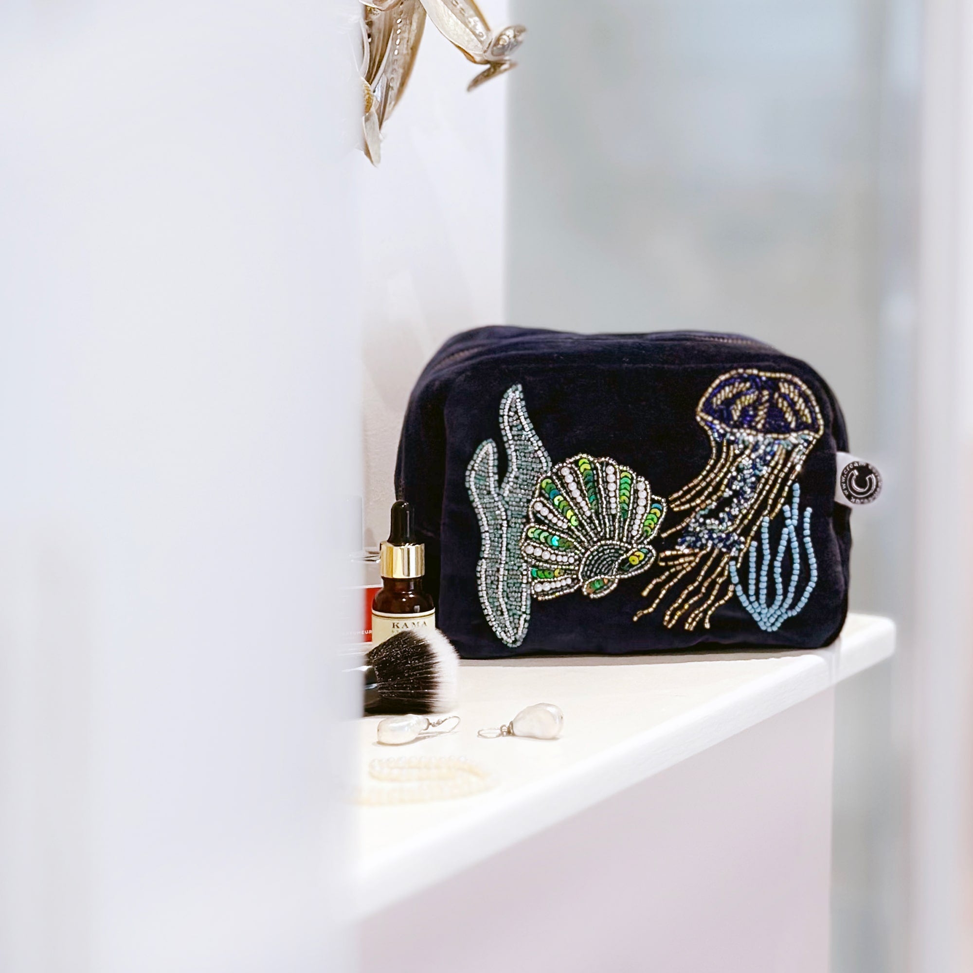 Beaded cosmetic bag in navy velvet with underwater scene of jellyfish, scallop shell and seaweed sitting on a bathroom shelf with some cosmetics around it.