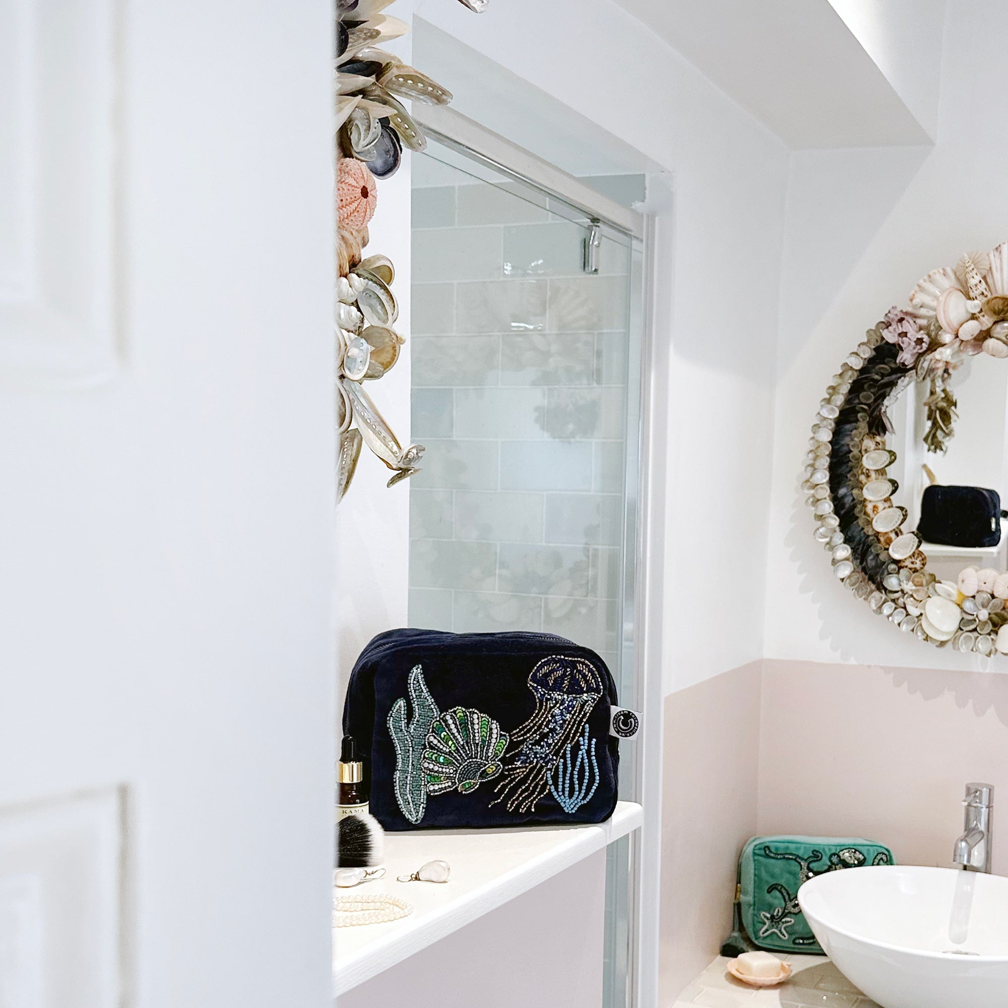 Beaded cosmetic bag in navy velvet with underwater scene of jellyfish, scallop shell and seaweed sitting on a bathroom shelf with some cosmetics around it and the sink and a large shell mirror in the background.