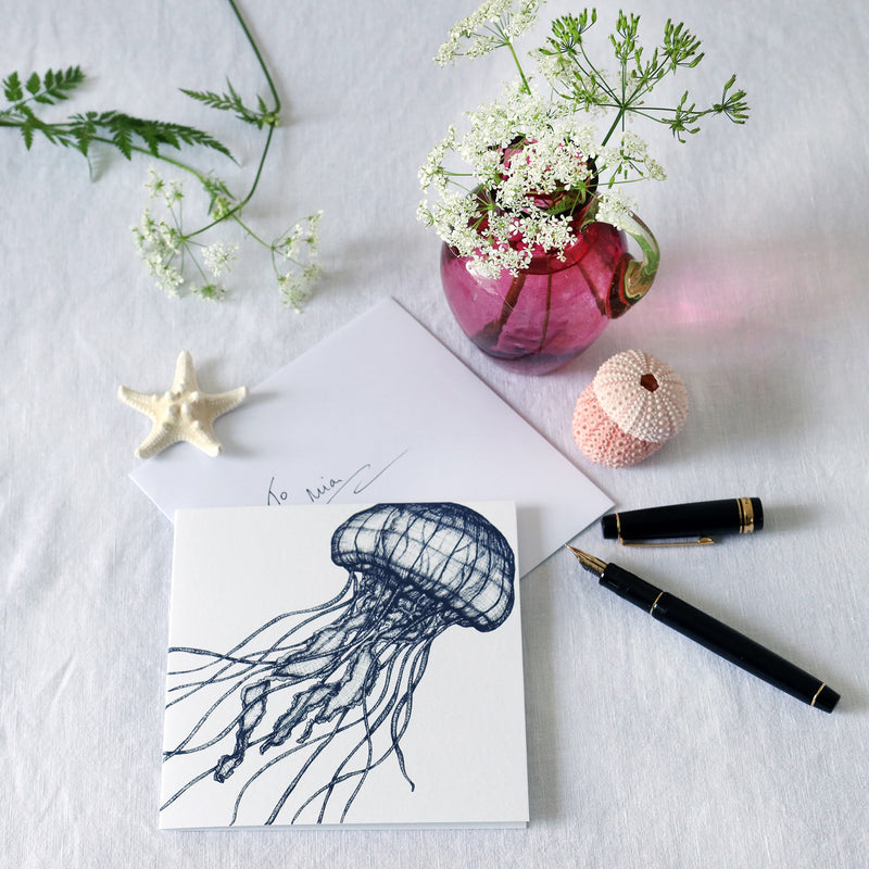 greeting card with illustrated navy jellyfish which looks like it's dancing on a white background lying on a white table cloth with a fountain pen, hand written envelope shells and a small cranberry glass jug with wild flowers in 