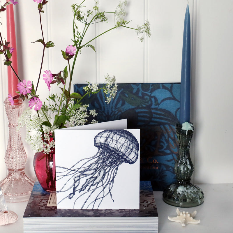 greeting card with illustrated navy jellyfish which looks like it's dancing on a white background on shelf with pink and blue candles in candlesticks and a small cranberry glass jug with wild flowers in