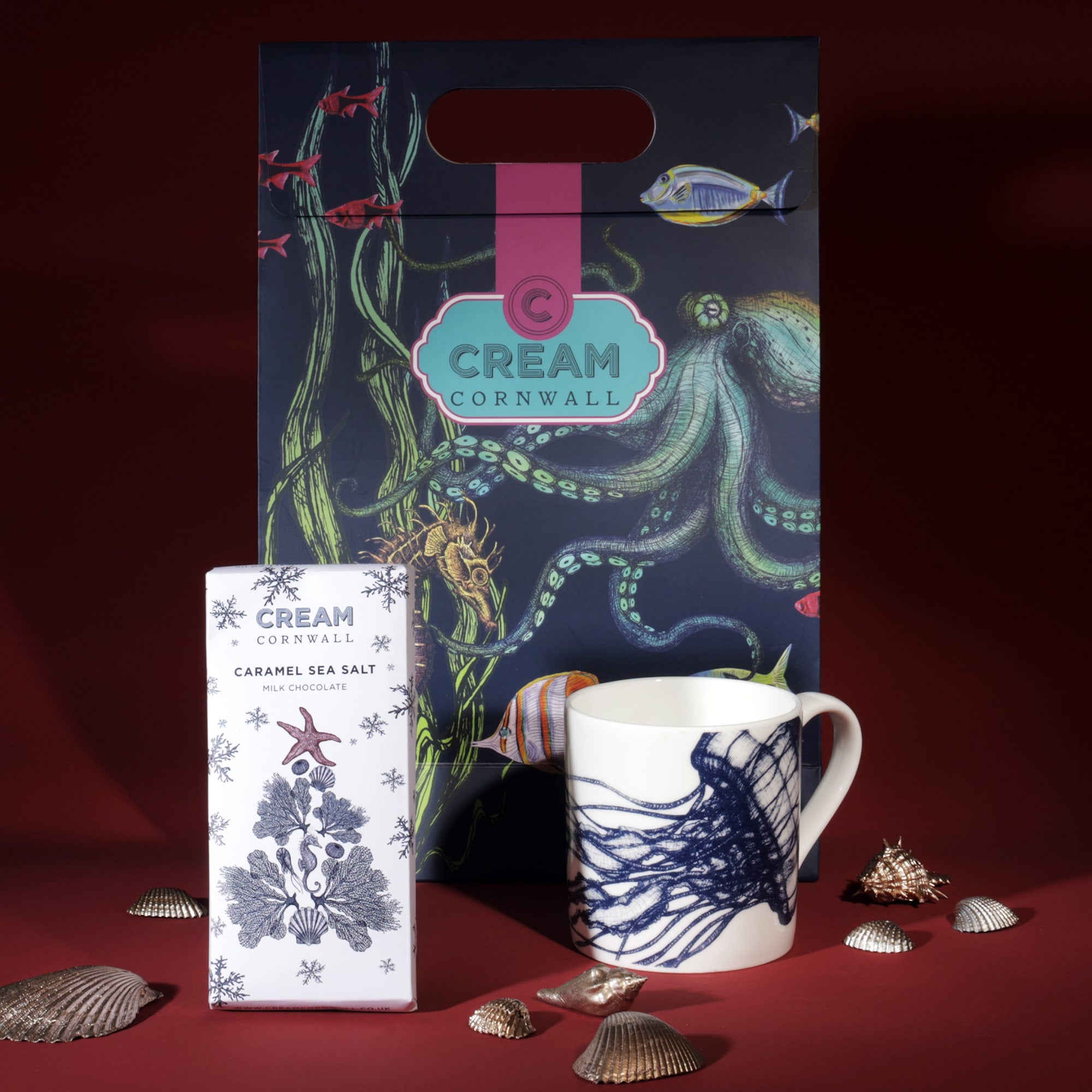 A Cream Cornwall gift set featuring a ceramic mug with a navy jellyfish design and a bar of caramel sea salt chocolate. The items are displayed with a colorful marine-themed gift bag, decorated with illustrations of fish, seaweed, and an octopus. Decorative seashells are scattered around the products, enhancing the coastal theme against a deep red background.