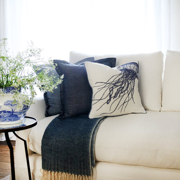 row of two blue cushions and a blue & white jellyfish cushion at the front, sitting on a navy throw on a white sofa with bright sunlight window behind and a large willow pattern jug filled with cow parsley