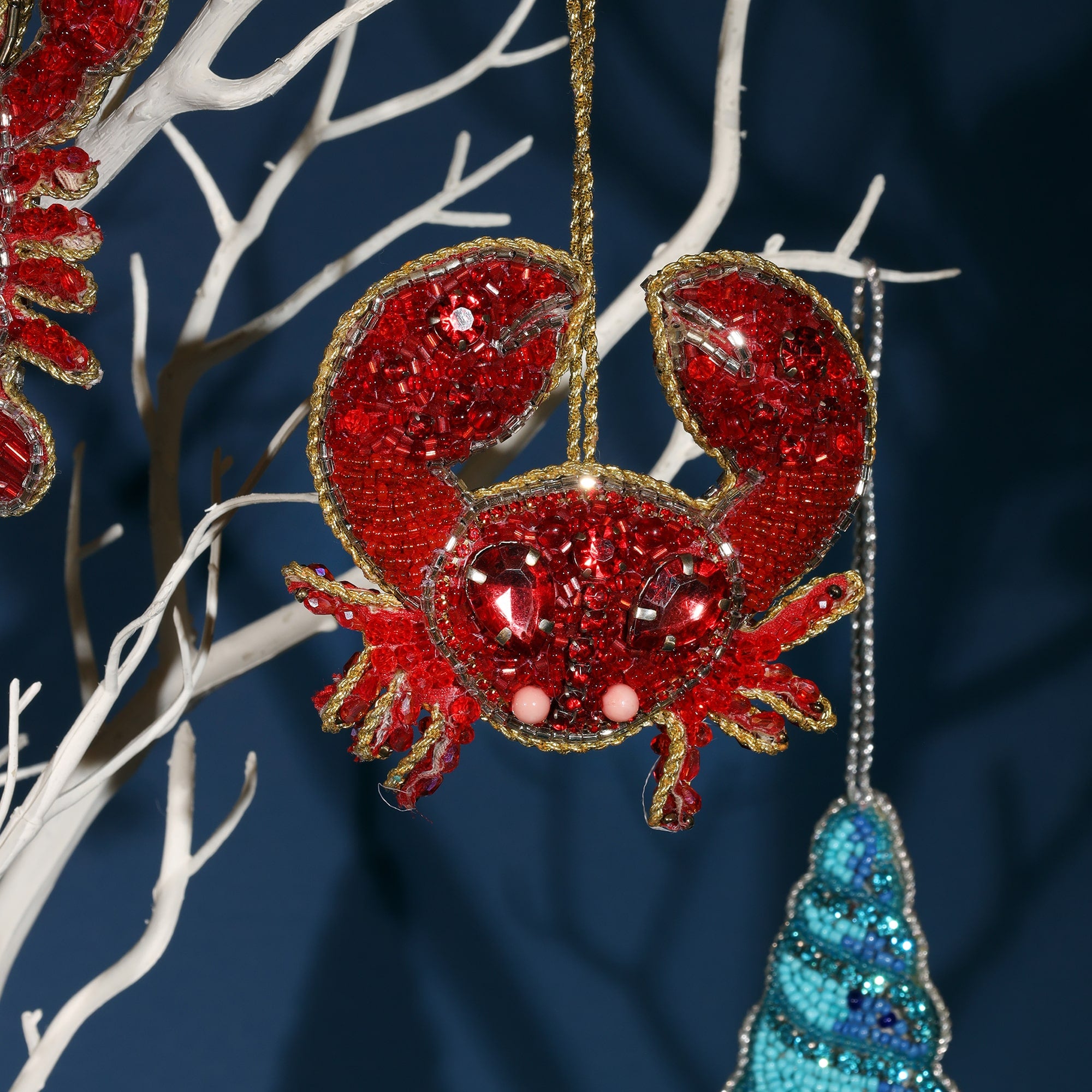 Close-up of a beaded red crab Christmas ornament. The crab is adorned with sparkling red beads and gemstones, with pink bead eyes and gold trim. It hangs from a gold string on a white decorative tree branch.