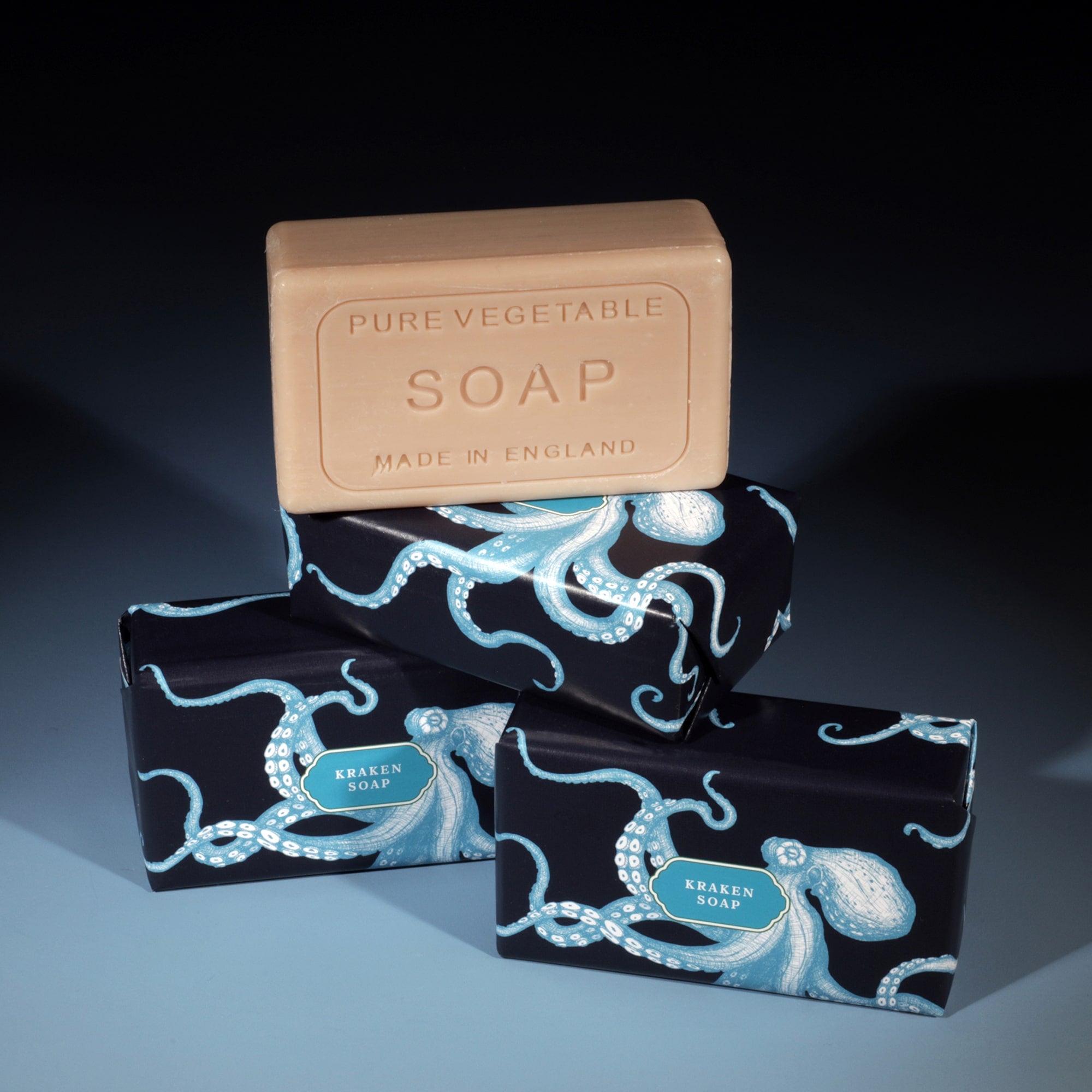 Three Kraken Soap bars are arranged against a dark background. Two bars are wrapped in black and blue paper adorned with octopus illustrations, while one bar of unwrapped pure vegetable soap, marked 'Made in England,' rests on top.