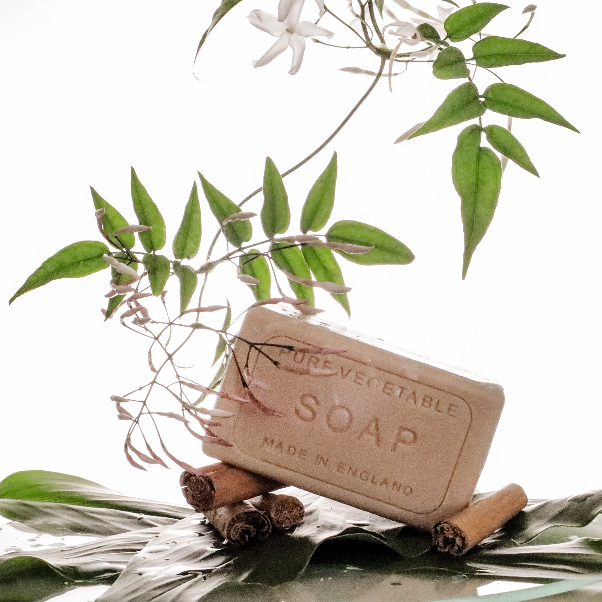 A bar of pure vegetable soap, engraved with 'Made in England,' is artfully displayed on two cinnamon sticks. Surrounding it are delicate jasmine flowers and green leaves, arranged on a glossy tropical leaf against a white background.