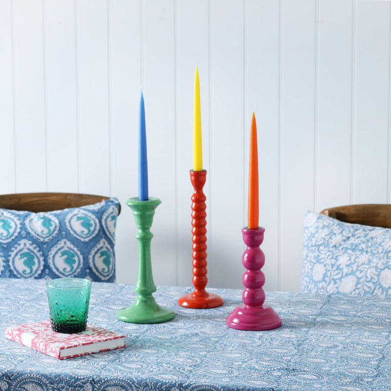 Three lacquered candle holders in various colours with contrasting candles in.They are placed on our Samudra collection table linens,also on the table is a notebook and a glass.In the background are a couple of chairs with hand blocked painted cushions
