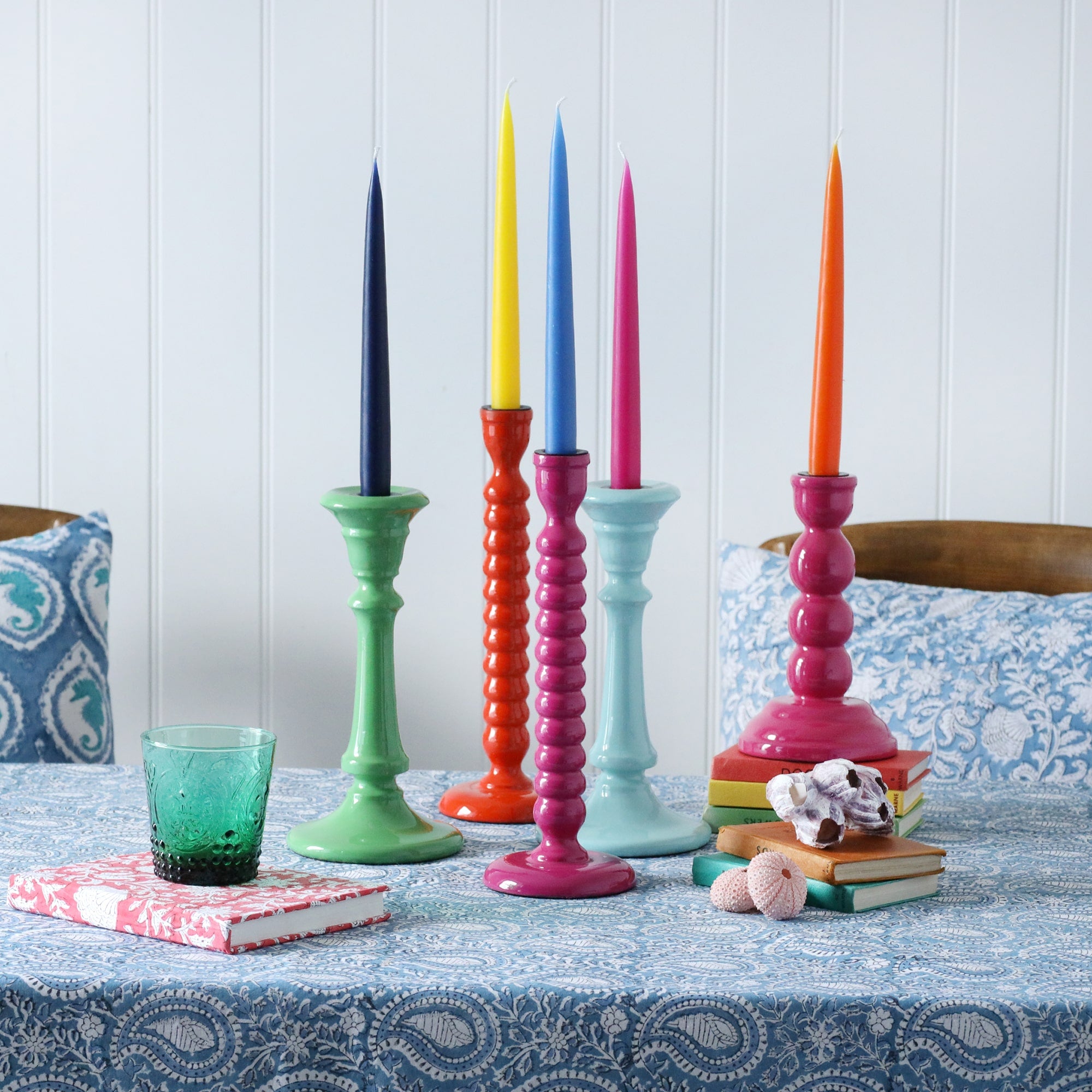 Various candle holders placed on our Samudra collection linen.All of the candle holders have contrasting candles.Also on the table are notebooks and shells,in the background are some of our matching Samudra cushions