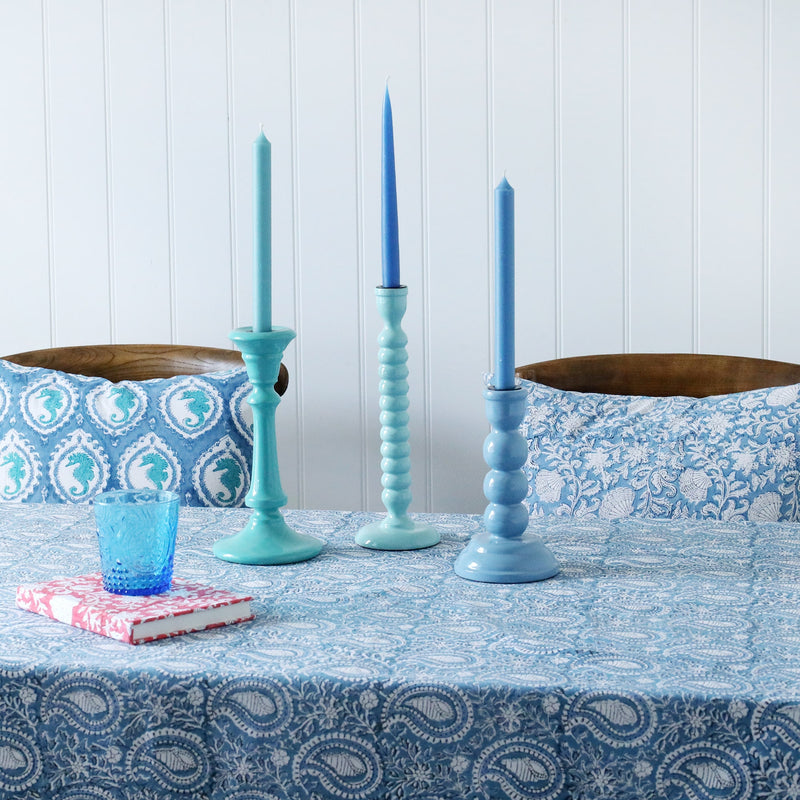Three lacquered candle holders in various colours with matching candles in.They are placed on our Samudra collection table linens,also on the table is a notebook and a glass.In the background are a couple of chairs with hand blocked painted cushions