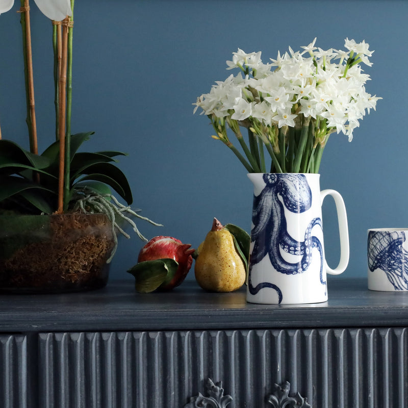 A white china jug with navy illustrated octopus on, filled with dainty spring daffodils sitting on a blue Gustavian set of reeded drawers. There are glazed porcelain fruits, and orchid and a pot with a jellyfish illustration sitting next to it.