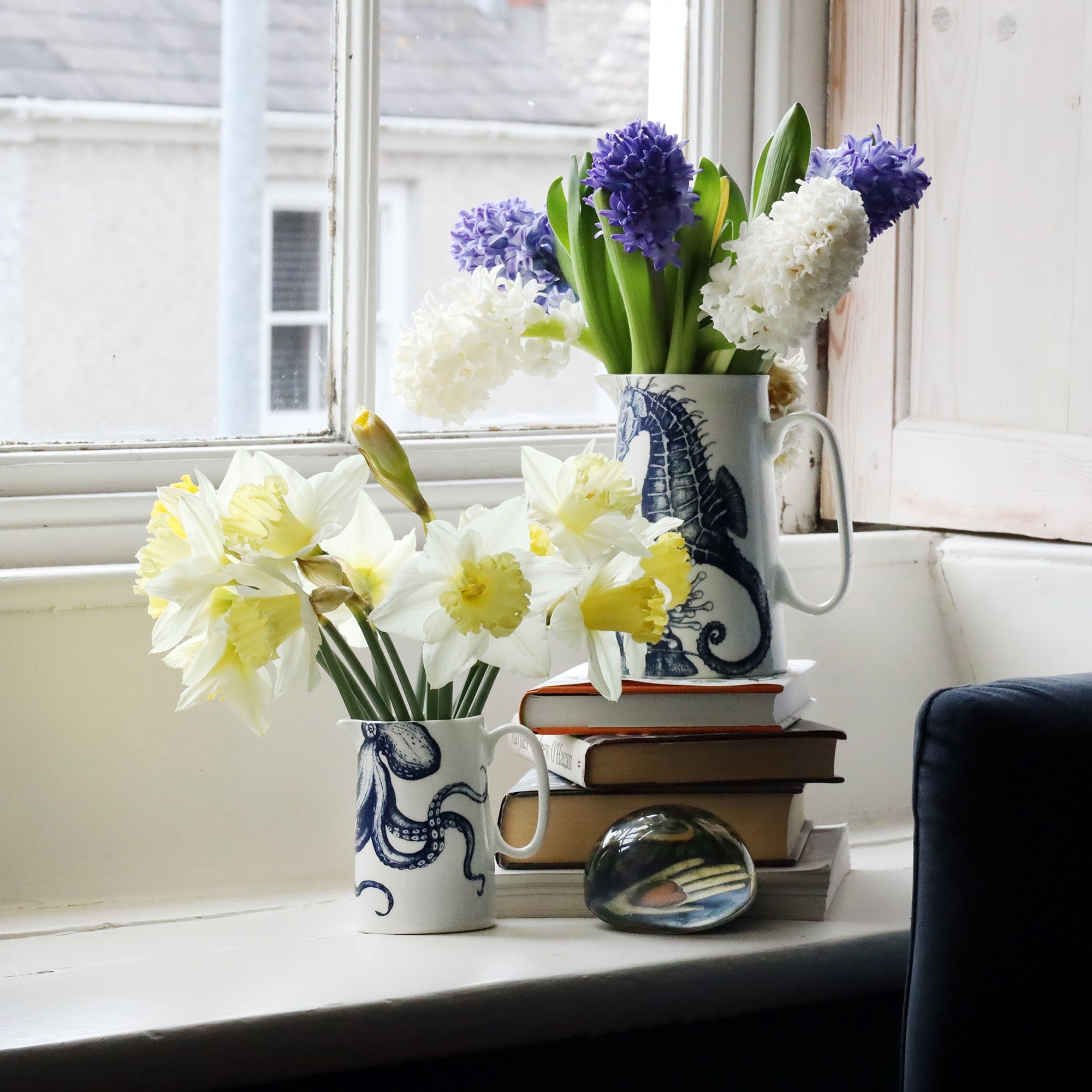 A painted wooden window seat with a pile of books and 2 jugs filled with spring flowers. One jug has an illustration of a seahorse and the other an octopus, both of which are in navy blue.