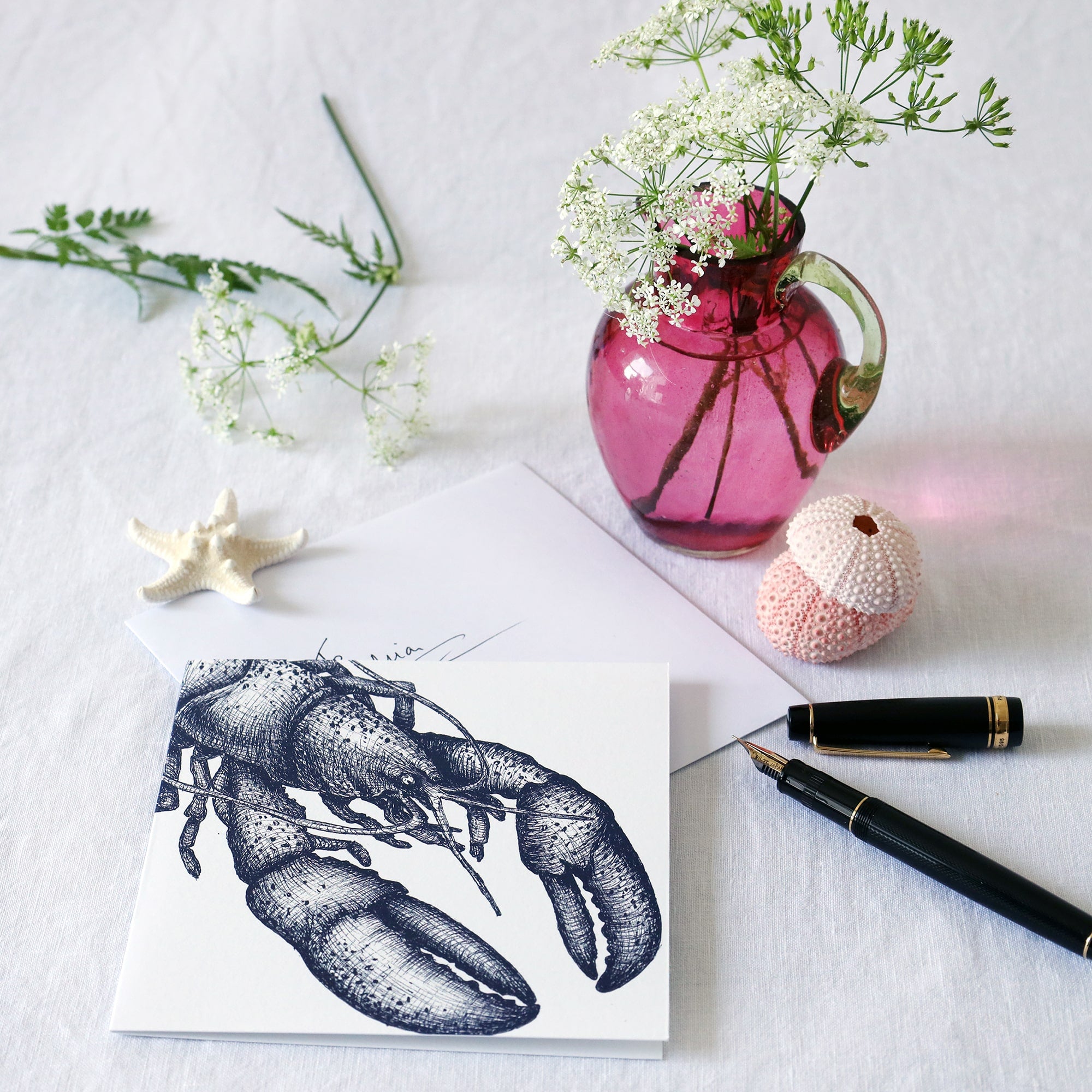 greeting card with navy illustrated lobster on white background lying on a white table cloth with a fountain pen, hand written envelope shells and a small cranberry glass jug with wild flowers in 