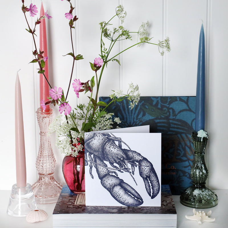 greeting card with navy illustrated lobster on white background on shelf with pink and blue candles in candlesticks and a small cranberry glass jug with wild flowers in