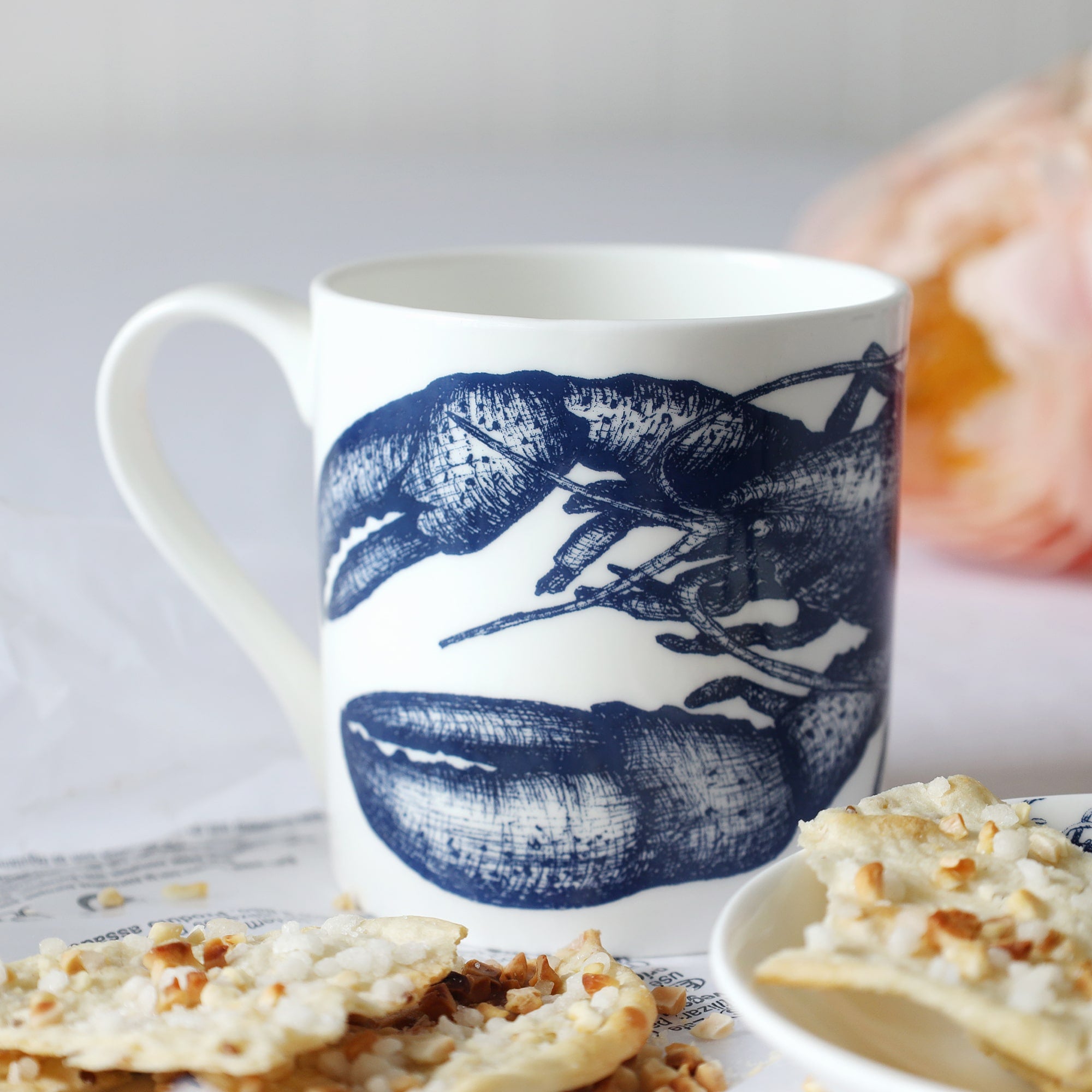 A white bone china mug with a beautiful dark blue illustrated lobster that runs around the middle sitting on a table with biscuits in front and out of focus flowers in the background.