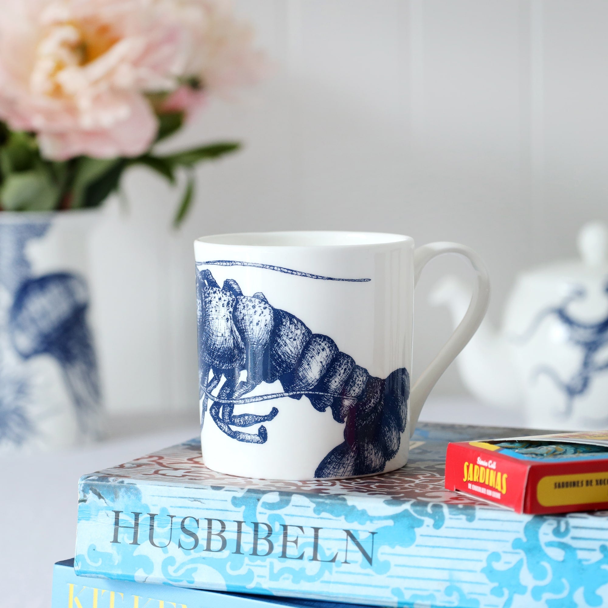 A white bone china mug with the tail of a blue illustrated lobster on it, sitting on a book with flowers & a teapot in the background.