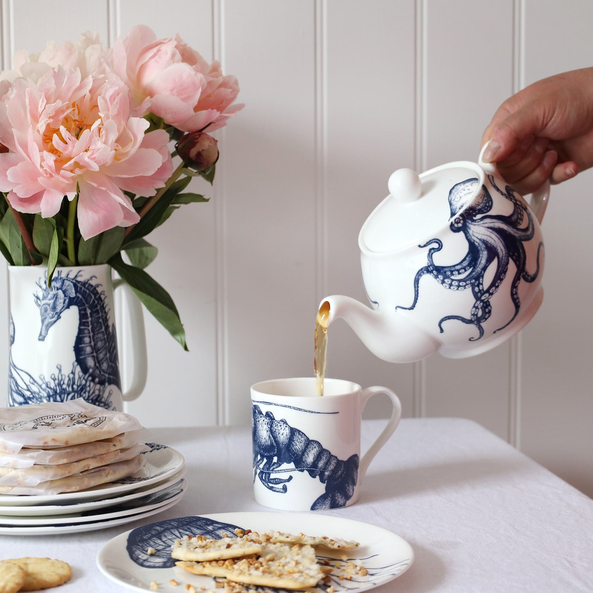 An informal table setting with blue & white coastal bone china on white linen and a shiplap background. There is an teapot with an octopus design pouring tea into a white mug that is decorated with a dark blue illustrated lobster tail.r 