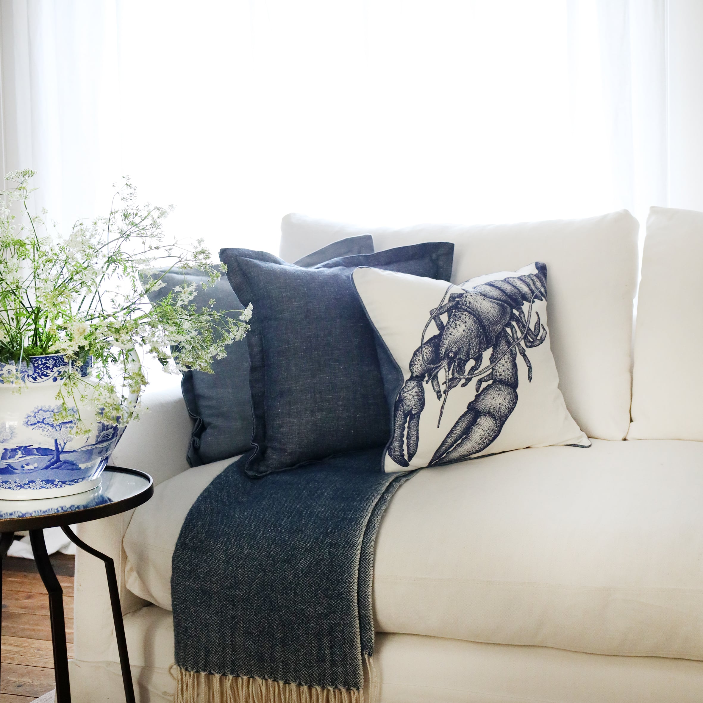 2 cushions in tones of blue with an illustrated lobster cushion at the front lined up on a white sofa with the sun streaming in from behind and a large willow pattern jug full of cow parsley