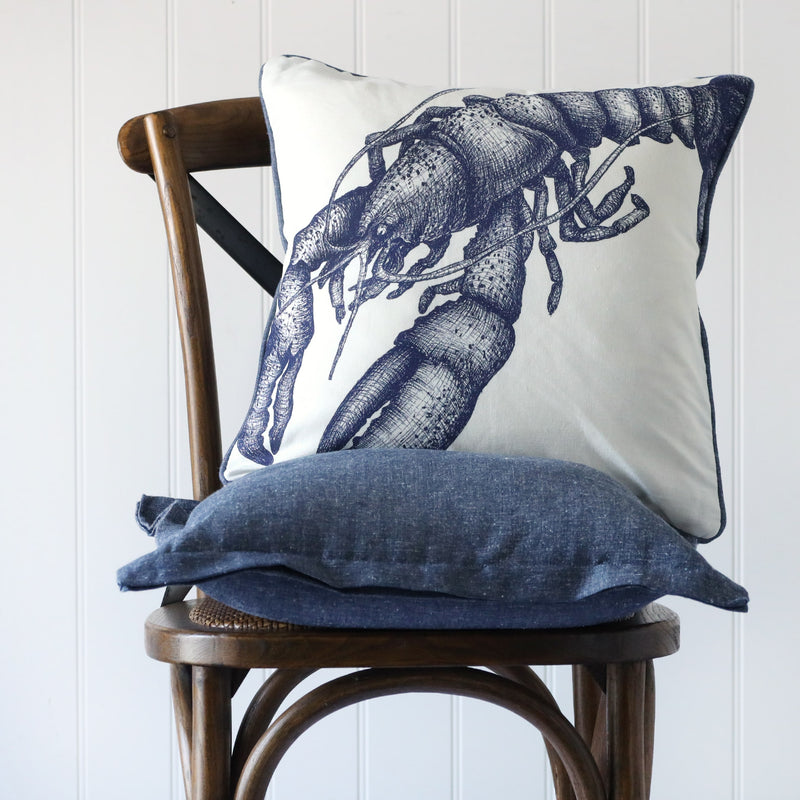 white cushion with navy illustrated lobster on the front, placed on a linen cushion on a wooden chair