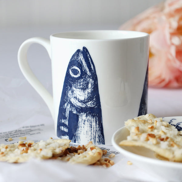 A close up of a white bone china mug with dark blue illustrated mackerel heads design coming up from the bottom of the mug. This is sitting on a white table cloth with a plate and biscuits and some flowers in the background.