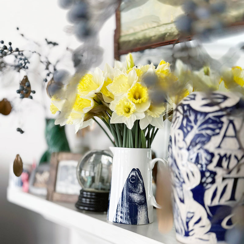 A white jug with blue mackerel heads facing upwards, filled with daffodils sitting on a mantlepiece with other vases and collected objets.