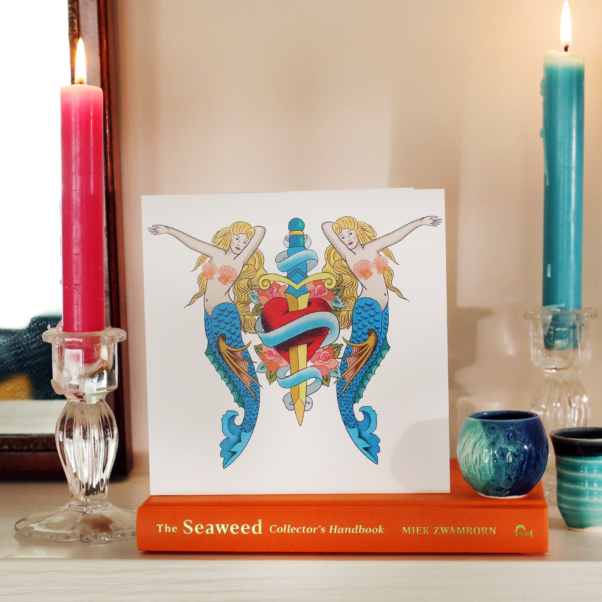 White card with illustrated mermaids & daggers tattoo inspired illustration, sitting on a book on a shelf with a candle lit each side and some small blue ceramic pots.