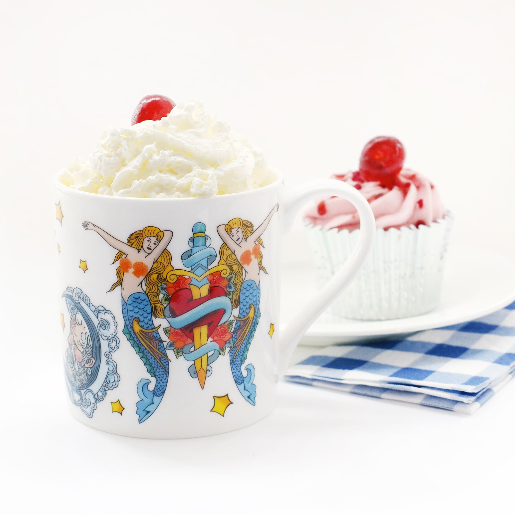 White mug decorated in a brightly coloured tattoo inspired design of mermaids and daggers. the mug is filled with whipped cream with a cherry on top. There is a pink cupcake with a cherry on top in the background and a blue checkered napkin.
