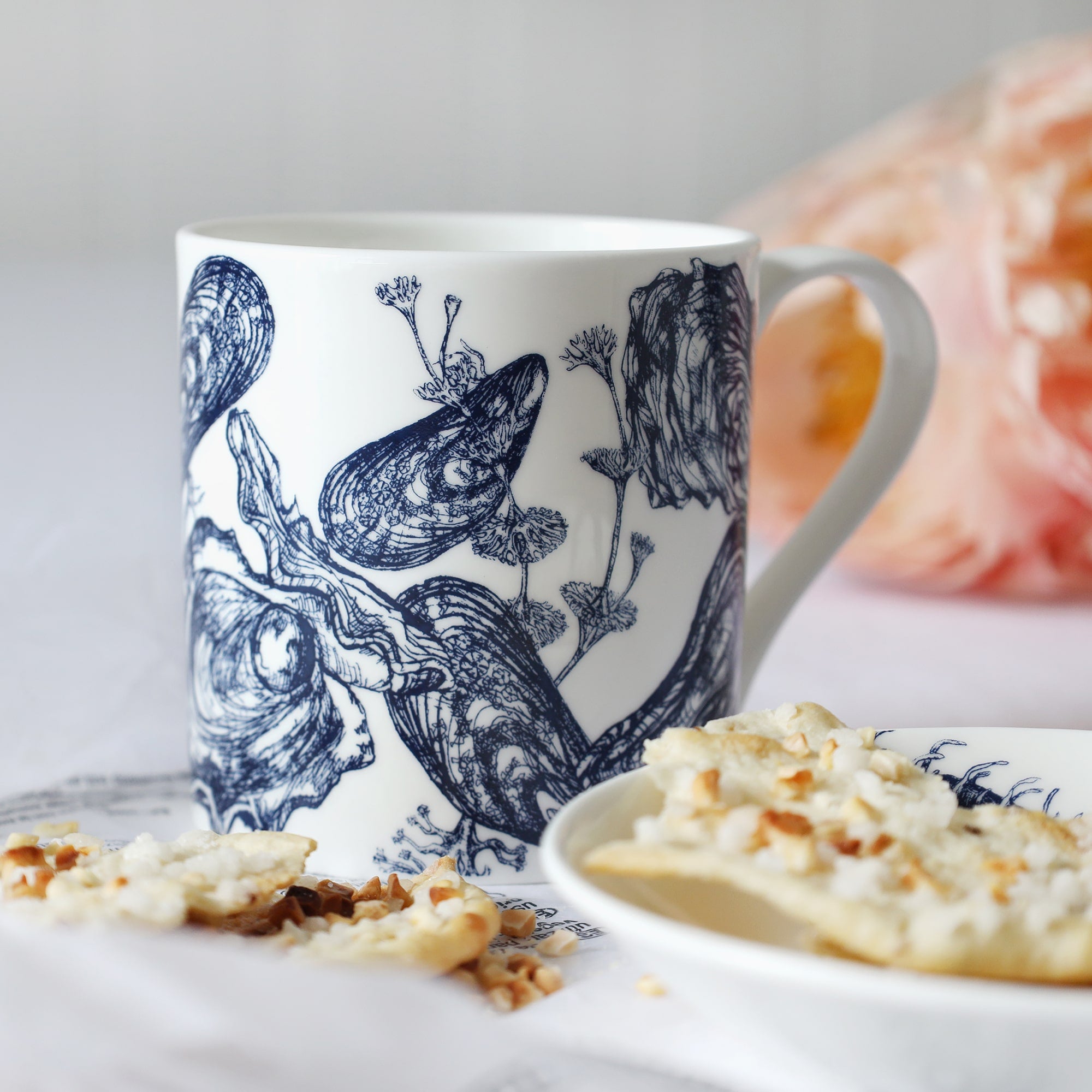 White mug with dark blue mussel & oyster shells illustration sitting on a table with a plate & biscuits and flowers in the background.