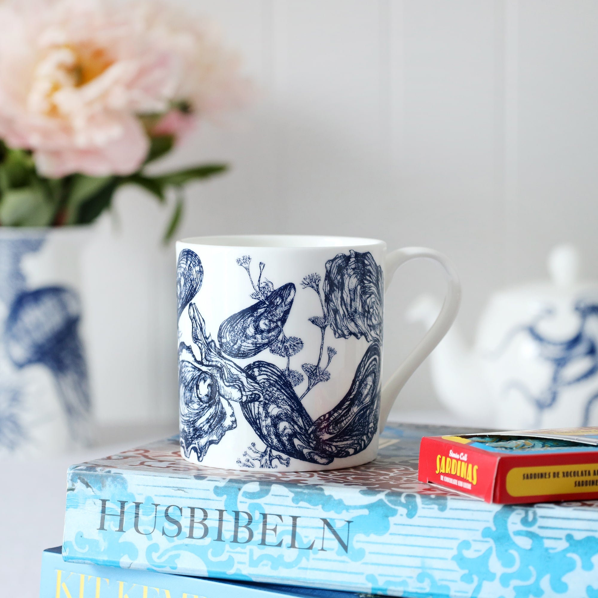 A close up of a white bone china mug with dark blue illustrated mussels & oyster design mug. This is sitting on a white table cloth with a plate and biscuits and some flowers in the background.