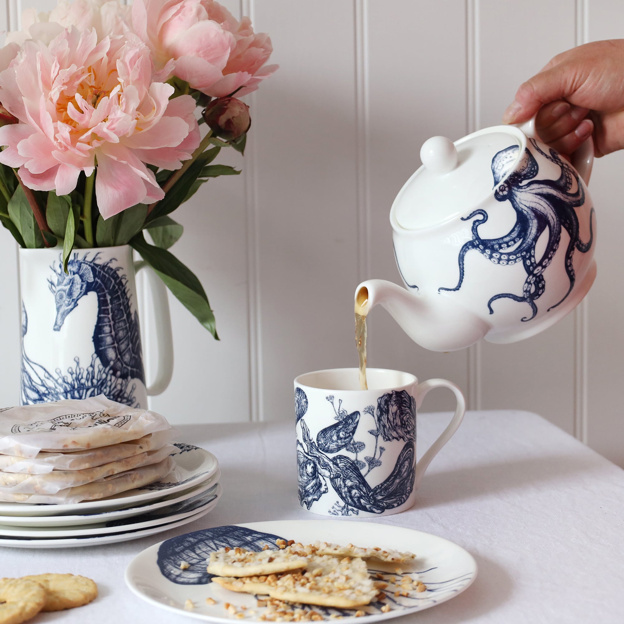 A blue & white informal; table setting set against a white shiplap wall and linen tablecloth. An octopus design adorns a teapot which is pouring tea into a white bone china mug decorated with a blue illustrated  mussel & oyster shell design..